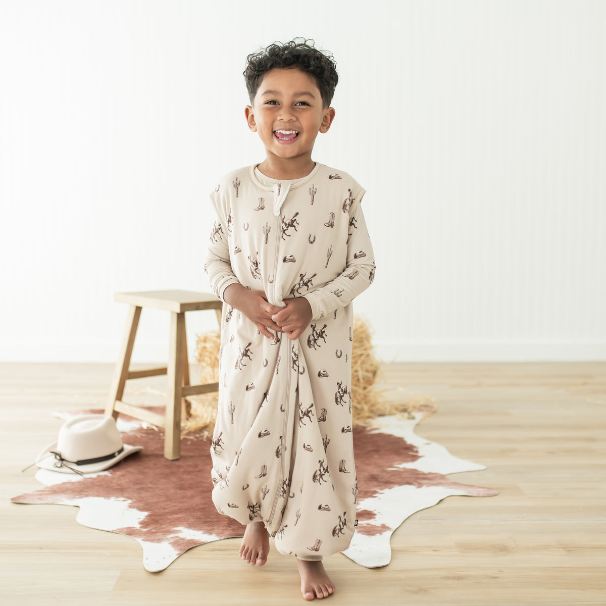 Smiling boy walking wearing the Sleep Bag Walker in Classic Cowboy 1.0 in front of a western inspired backdrop