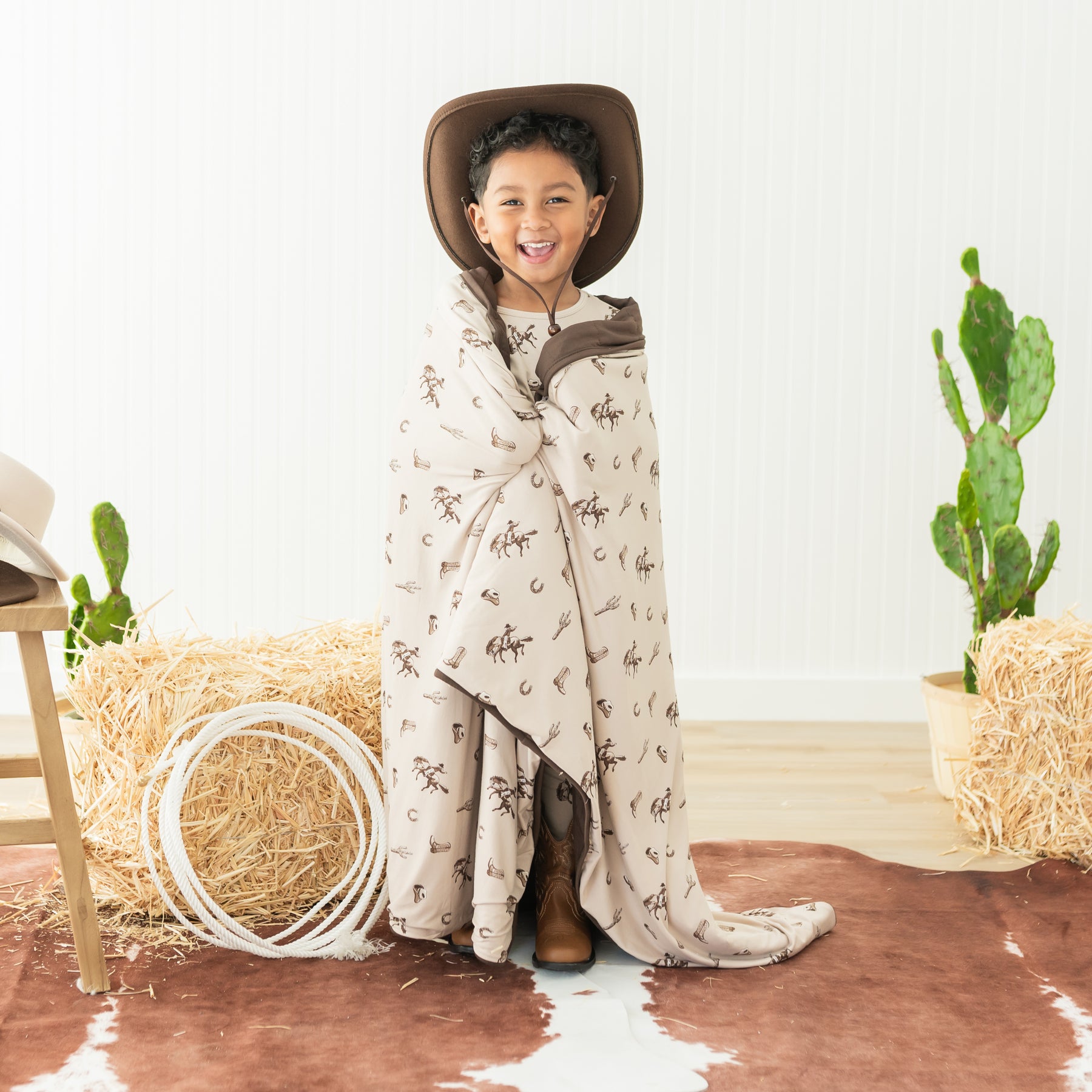 Young smiling boy standing in front of a western inspired back drop wearing a cowboy hat with the Toddler Blanket in Classic Cowboy 1.0 wrapped around him