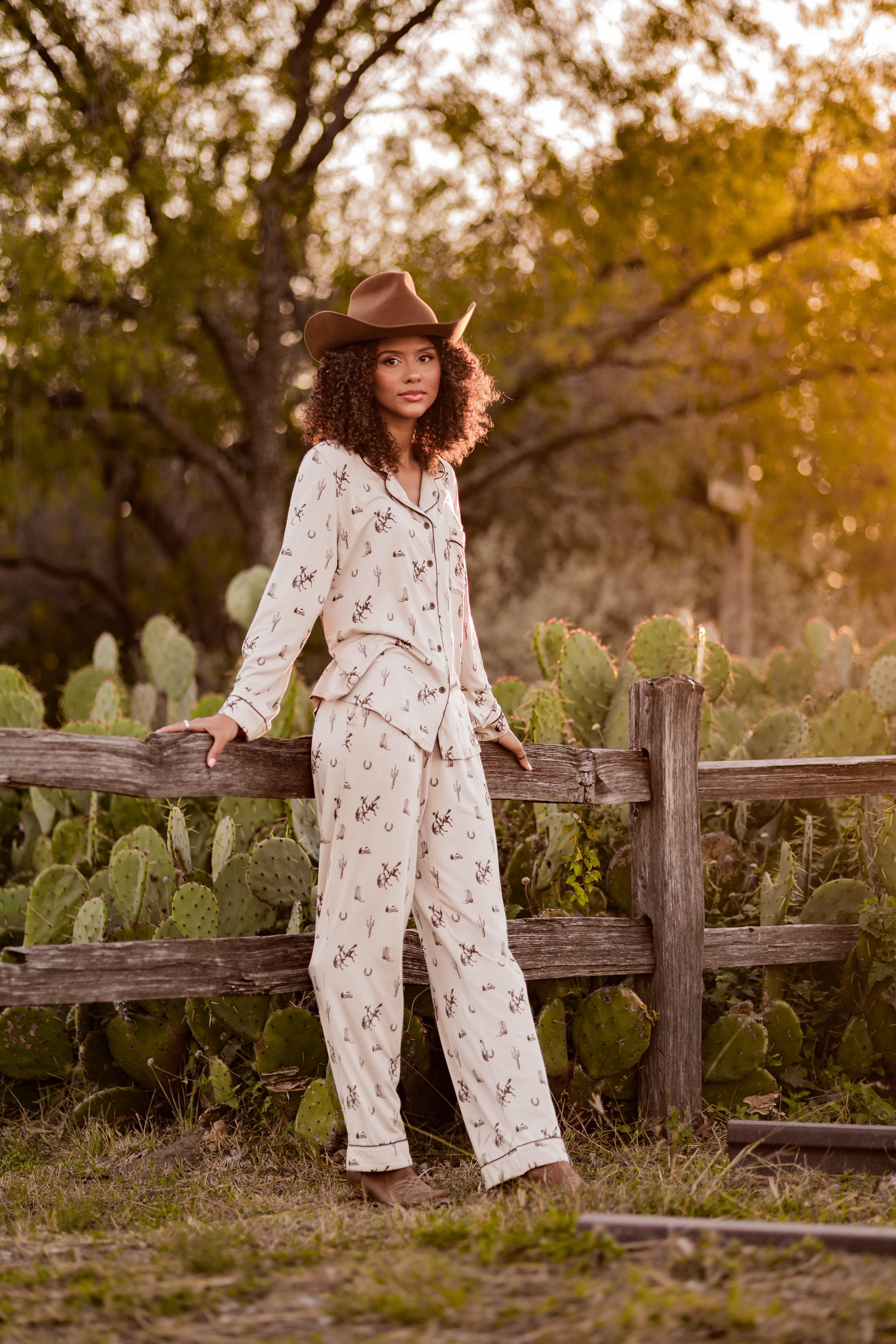 Female model wearing the Long-Sleeved Women's Pajama Set in Classic Cowboy leaning against a fence outside in front of cacti and trees