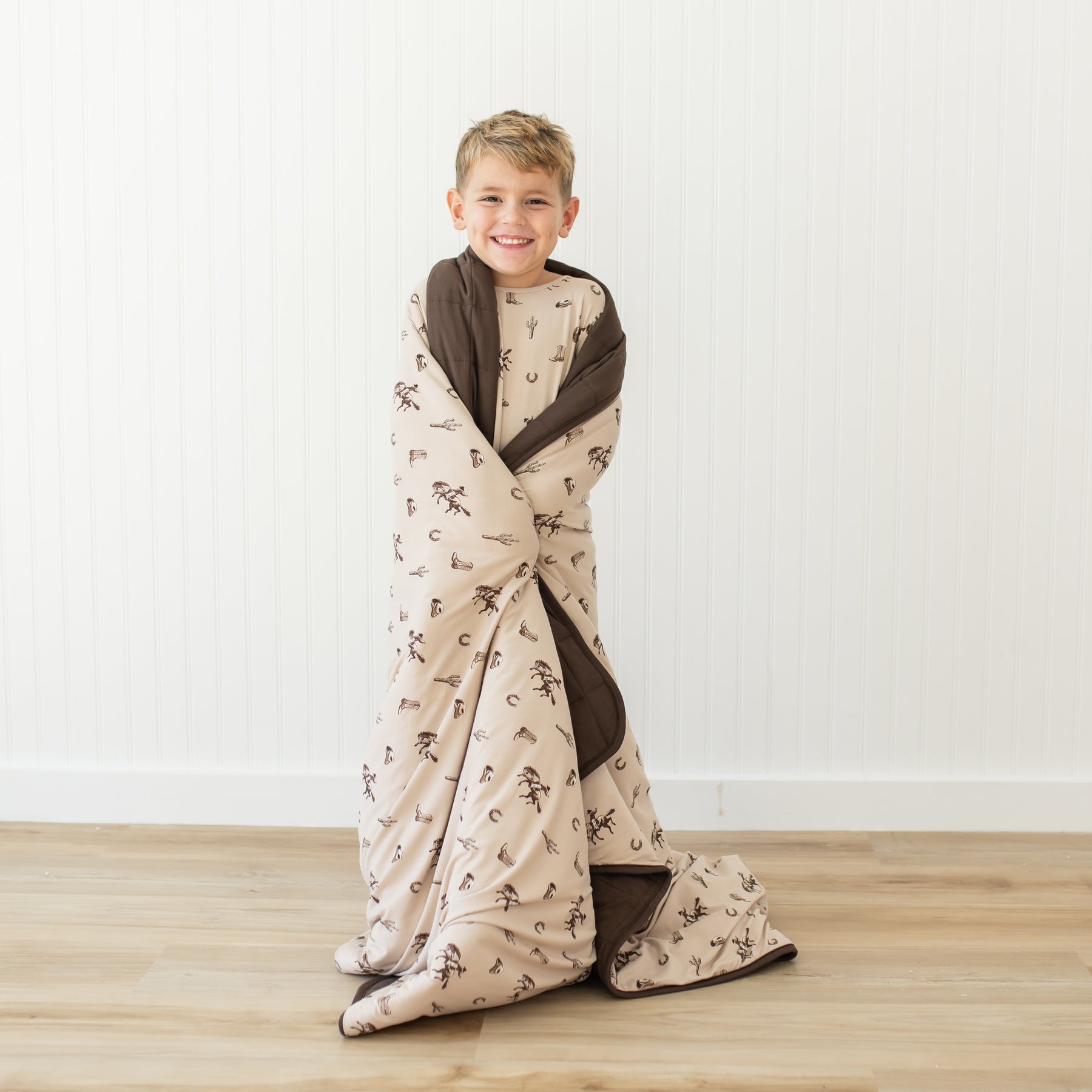 Smiling young boy standing in front of a white paneled wall with the Youth Blanket in Classic Cowboy 2.5 around his shoulders