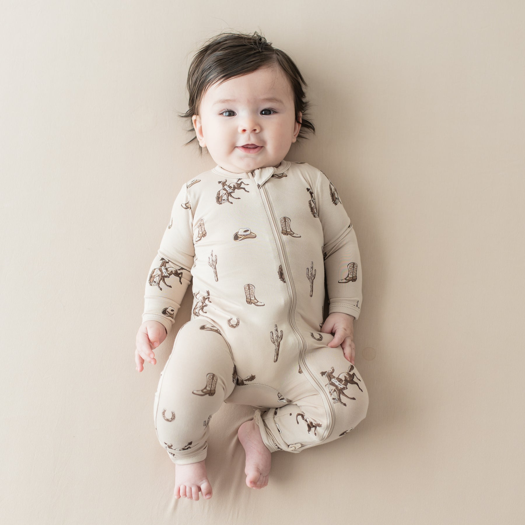 Infant laying on a light beige blanket wearing the Zippered Romper in Classic Cowboy