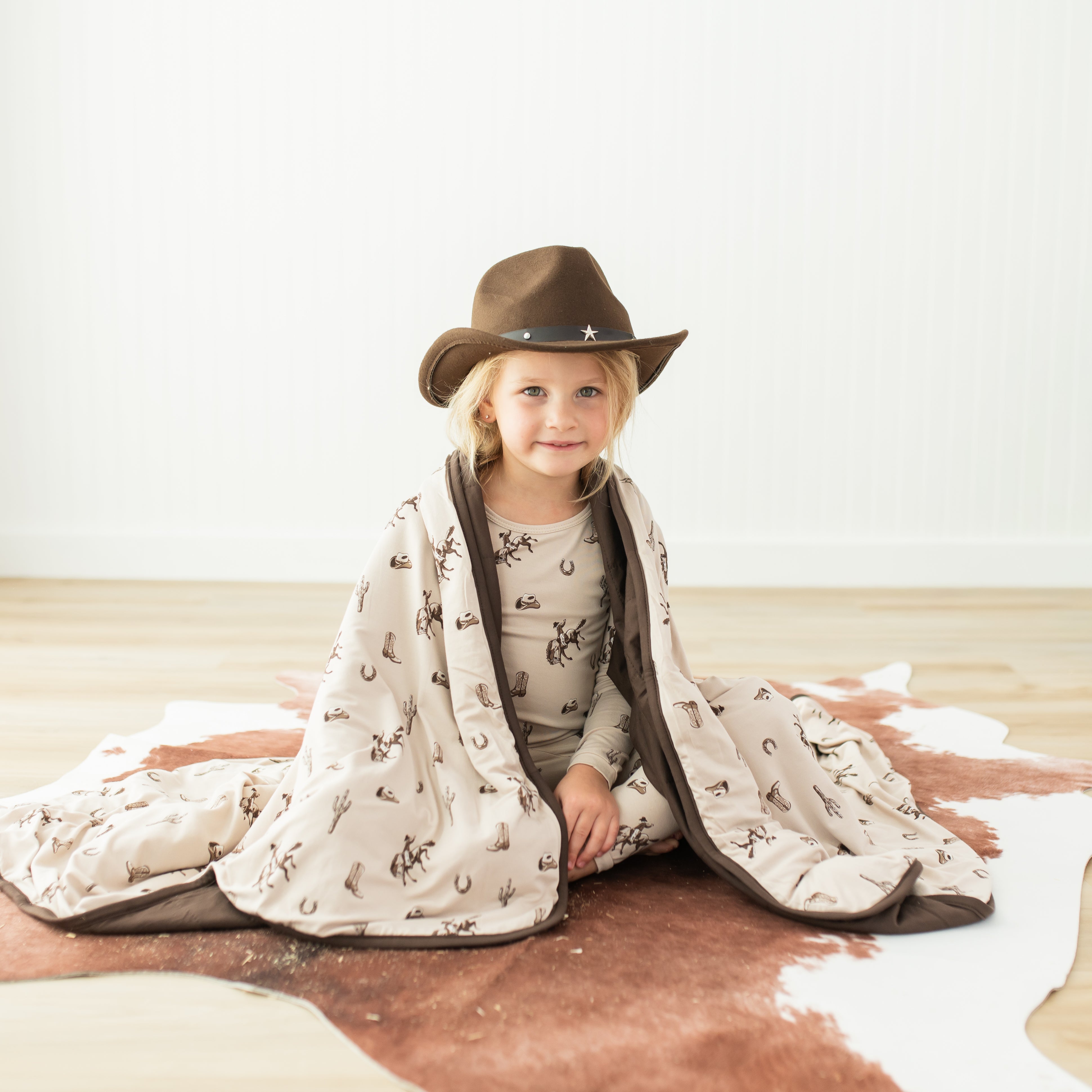 Young child sitting on the floor with the Youth Blanket in Classic Cowboy 2.5 wrapped around their shoulders wearing matching long sleeve pajamas and a brown cowboy hat