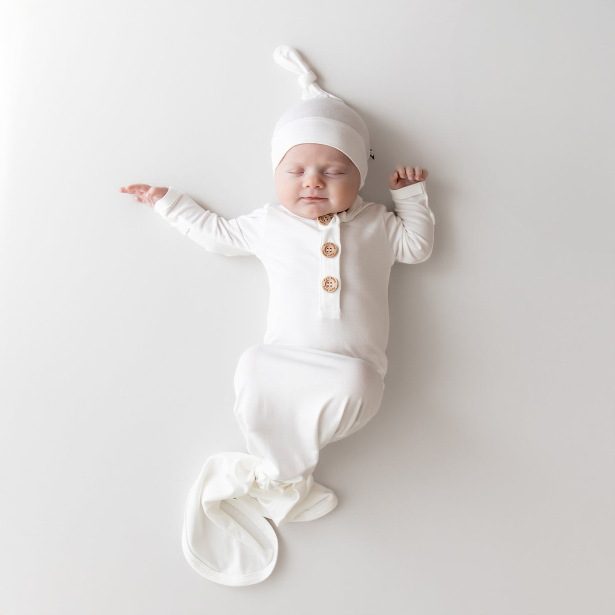 Newborn baby in a white outfit with buttons and a matching hat on a light gray background