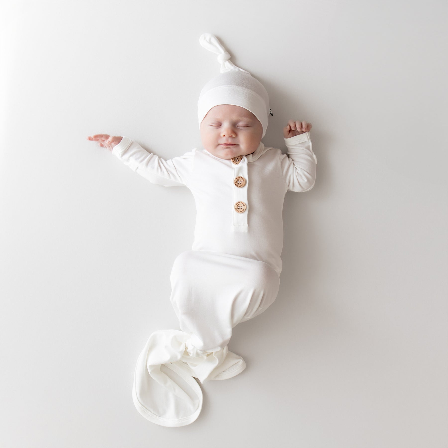 Newborn baby in a white outfit with buttons and a matching hat on a light gray background