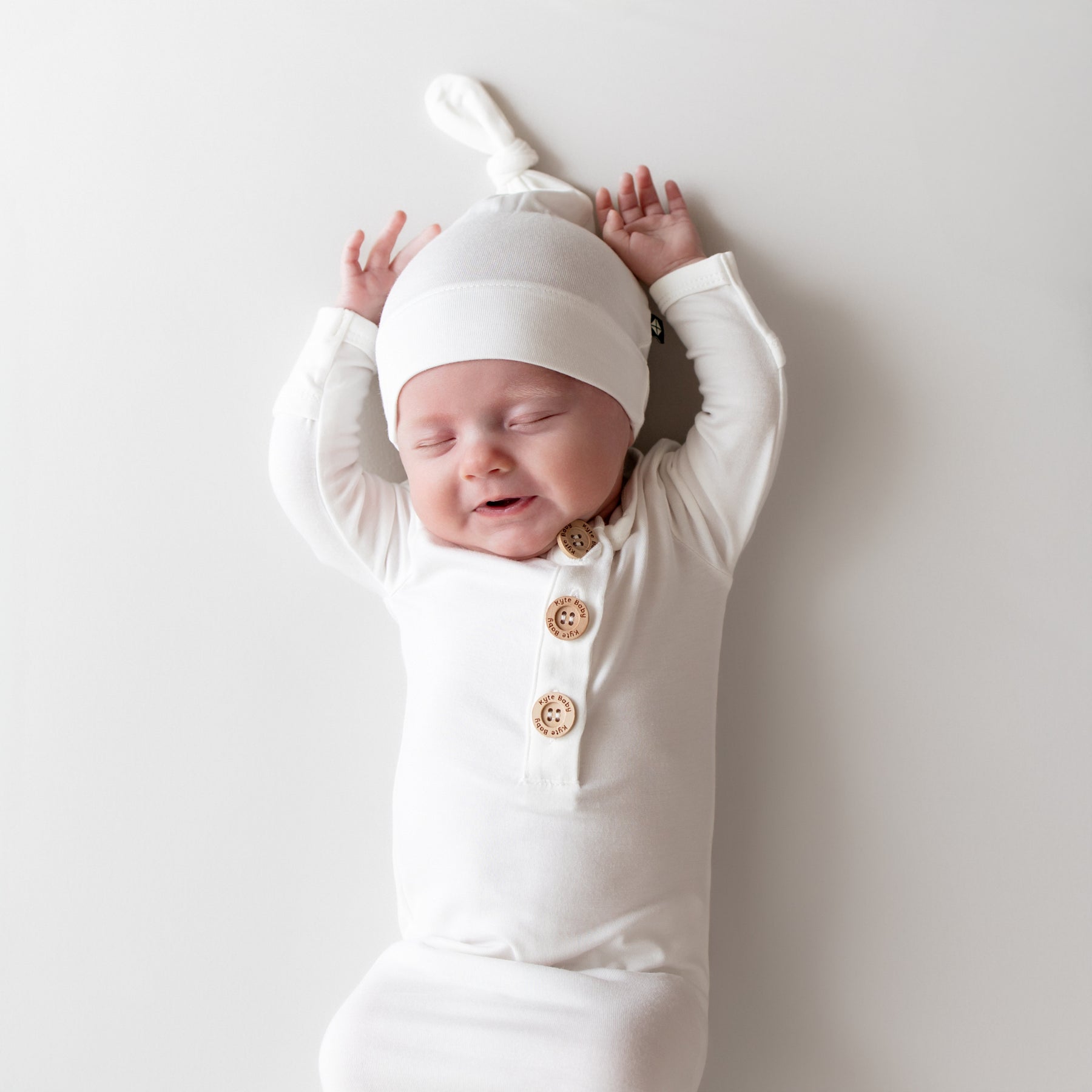 Newborn baby in a white outfit with buttons, wearing a matching white hat, against a plain background.