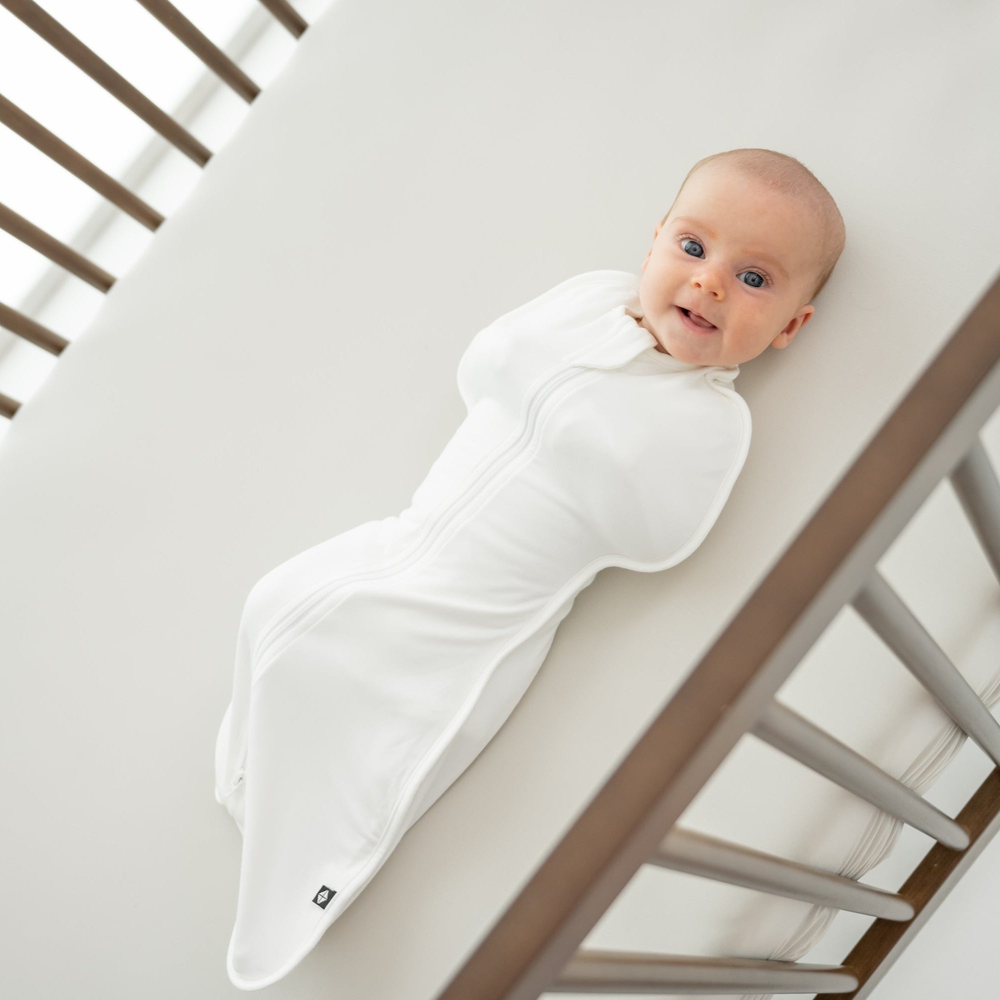 Baby in a wooden crib wearing a white swaddle bag 