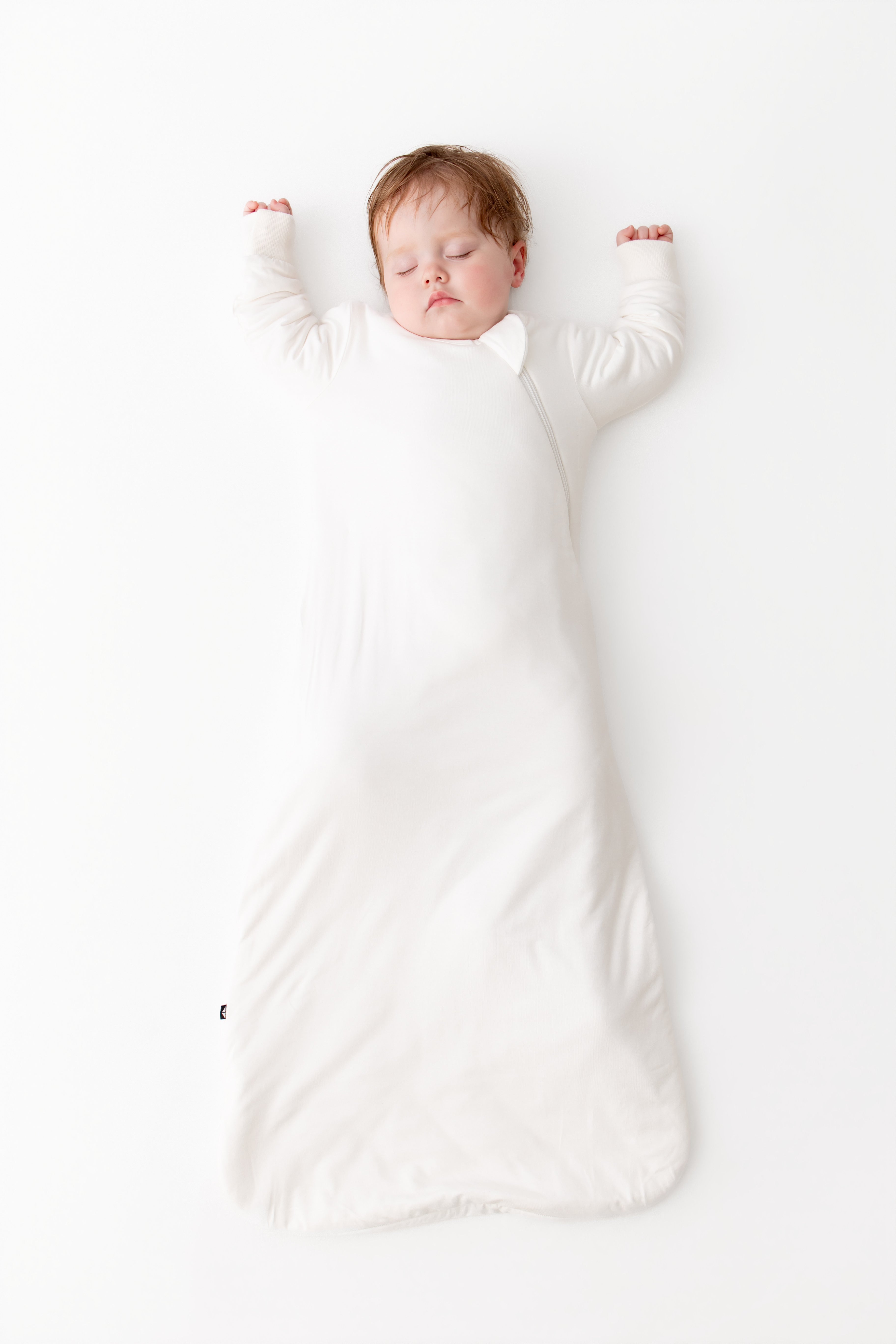 A sleeping newborn baby wearin a white long-sleeve sleep sack, lying on a clean white background.