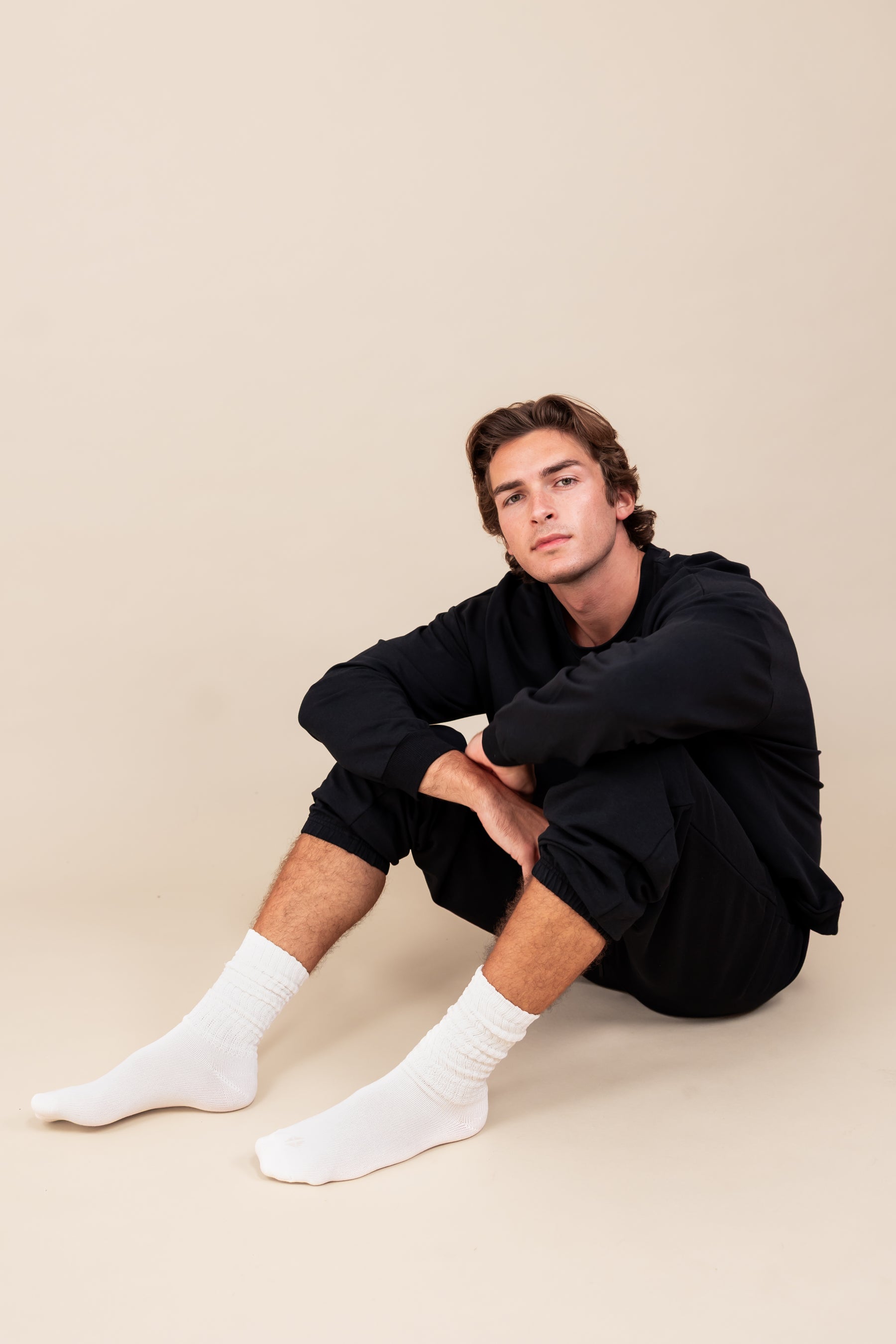 Modal sitting while wearing Adult Scrunch Socks in Cloud color on black jogger set on a beige background