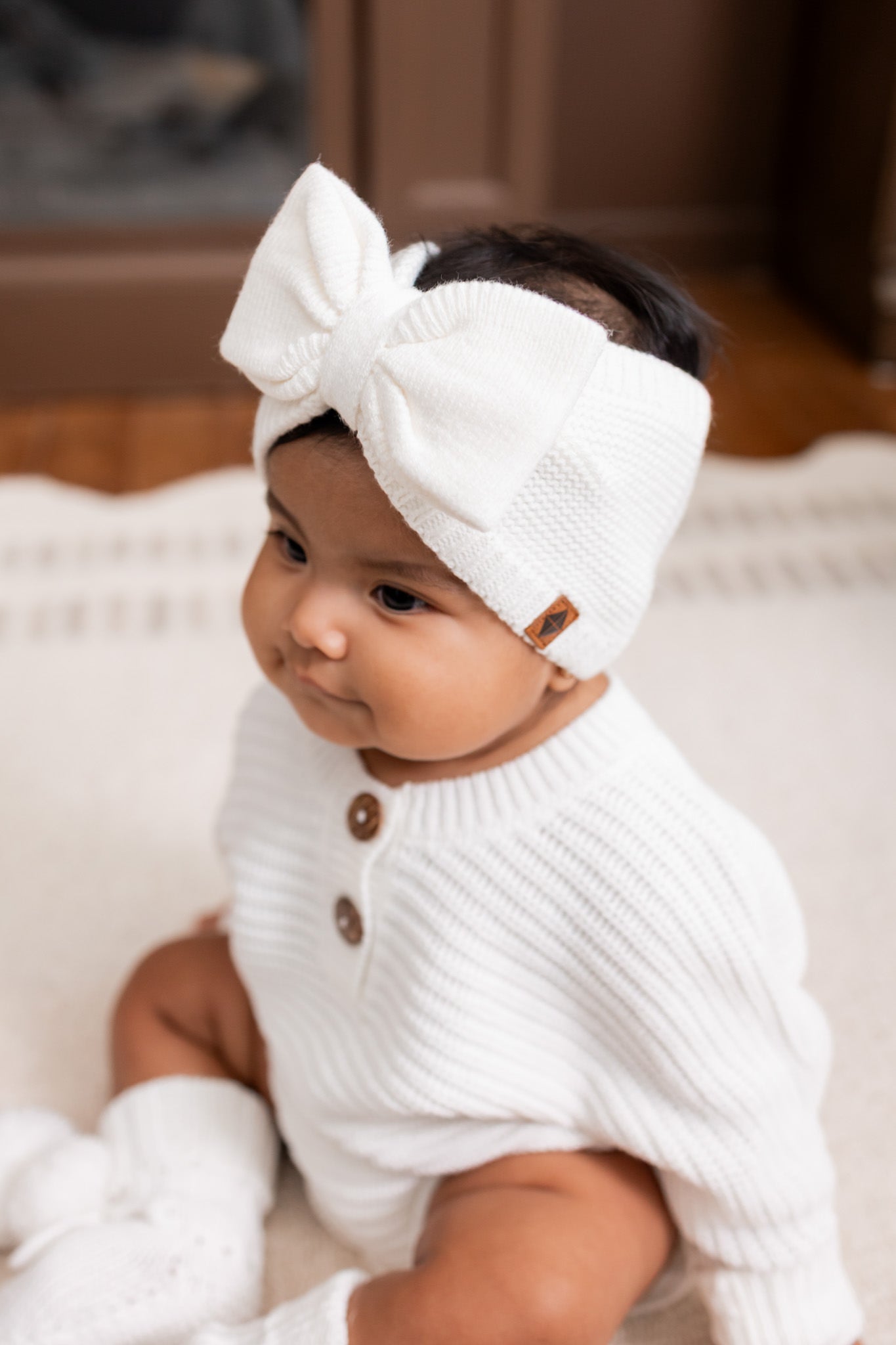 Baby wearing Chunky knit romper, Chunky knit bow headband and knit booties in cloud while sitting on a white carpet
