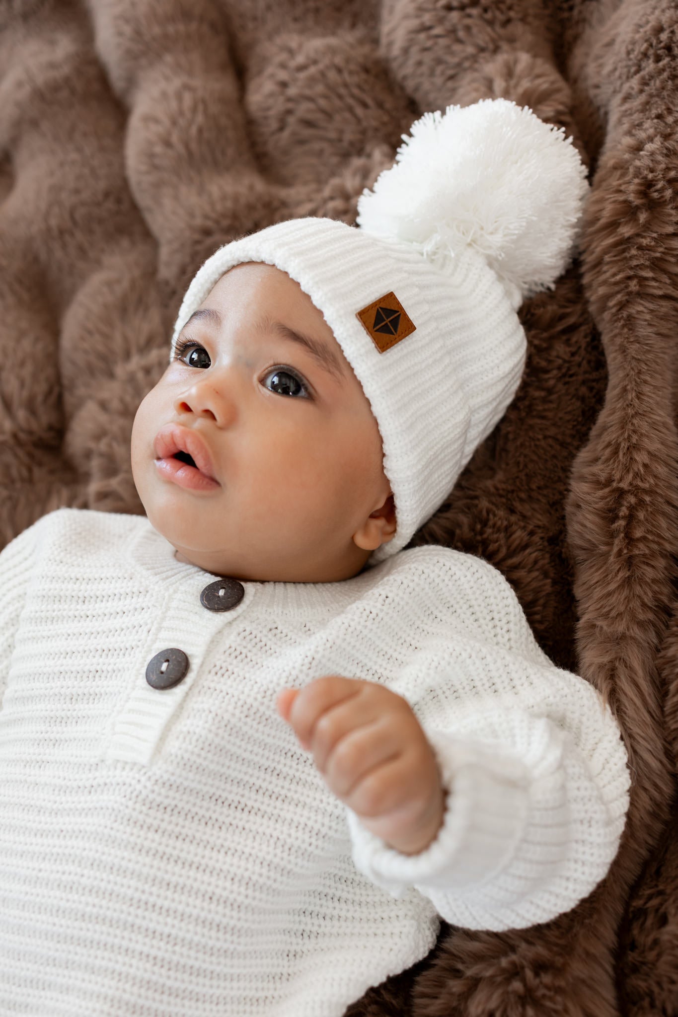 Baby wearing Chunky Knit Romper and chunky knit pom beanie in cloud lying on a brown fur blanket