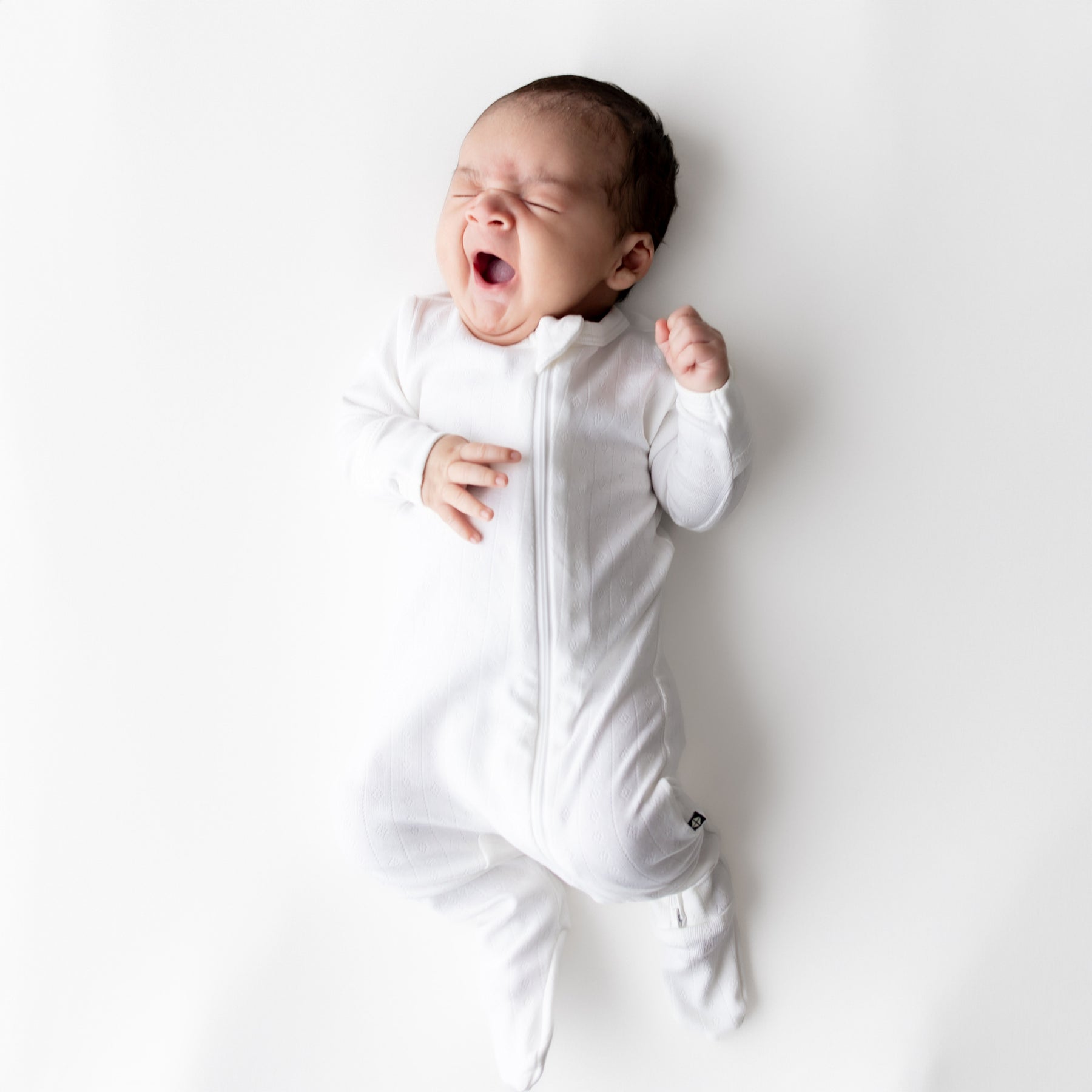 Yawning newborn wearing the Pointelle Zippered Footie in Cloud on a white surface