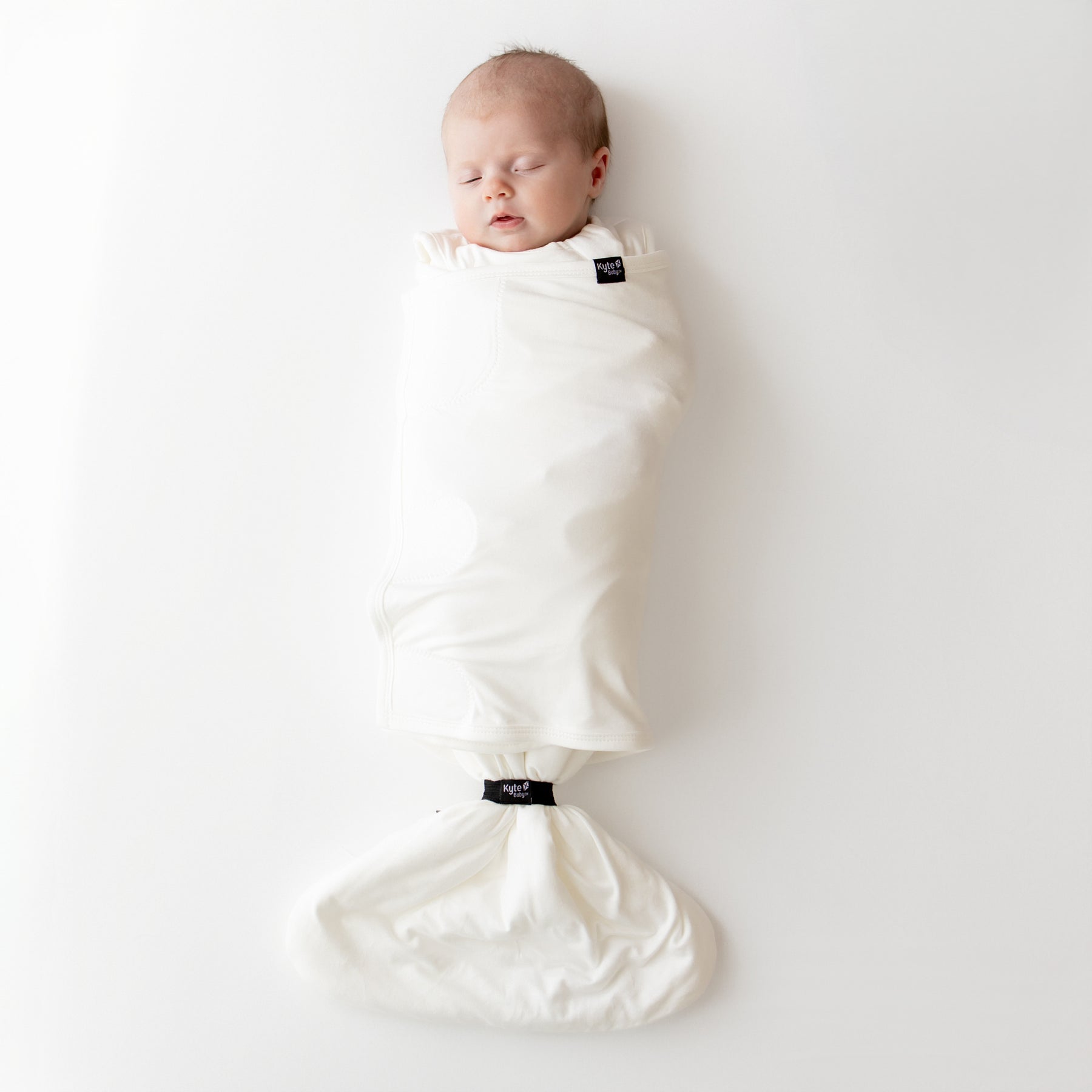 Newborn baby swaddled in a white sleep bag swaddler on a plain background