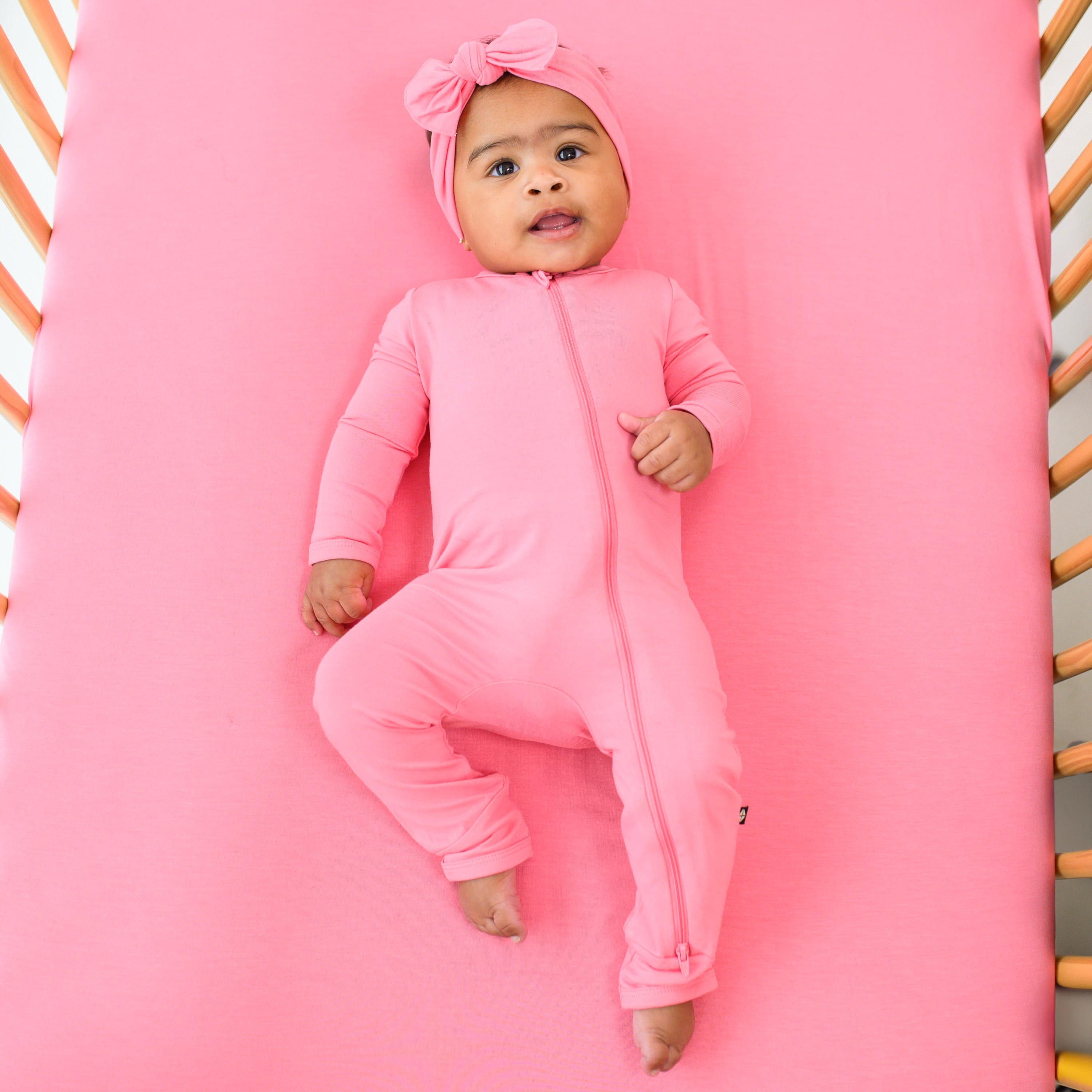 baby wearing zippered romper and bow in guava while laying on a crib mattress with guava crib sheet on