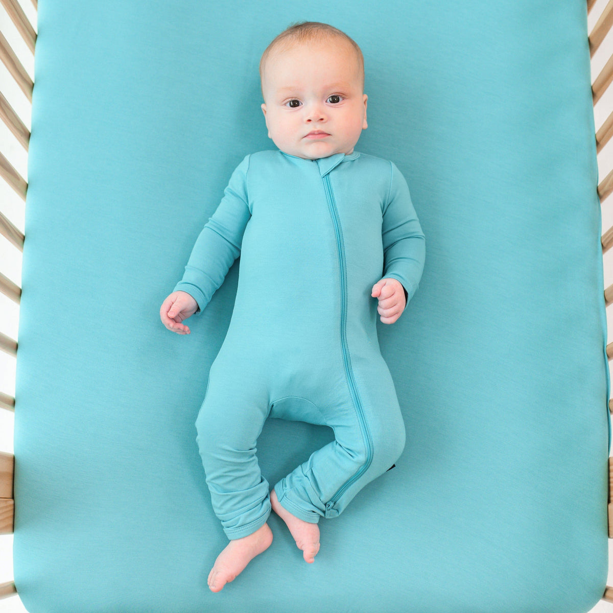 baby modeling zippered romper in makai while laying down in crib with a makai crib sheet on it