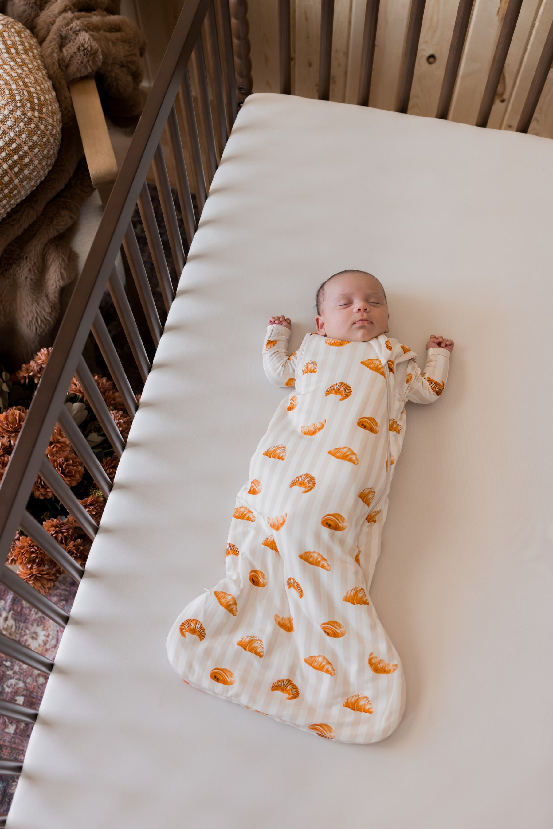 Baby sleeping in Sleep Bag with Croissant 1.0 print in a crib with beige sheet.