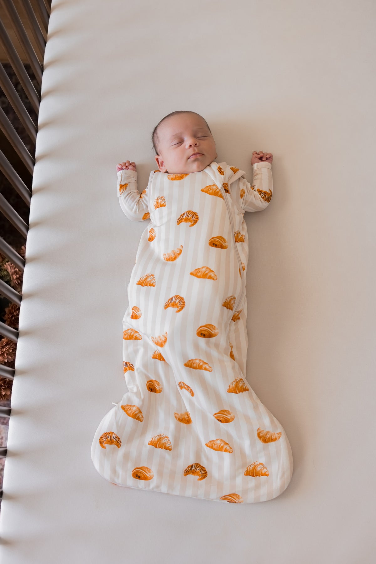 Baby sleeping in Sleep Bag with Croissant 1.0 print in a crib on beige crib sheet.