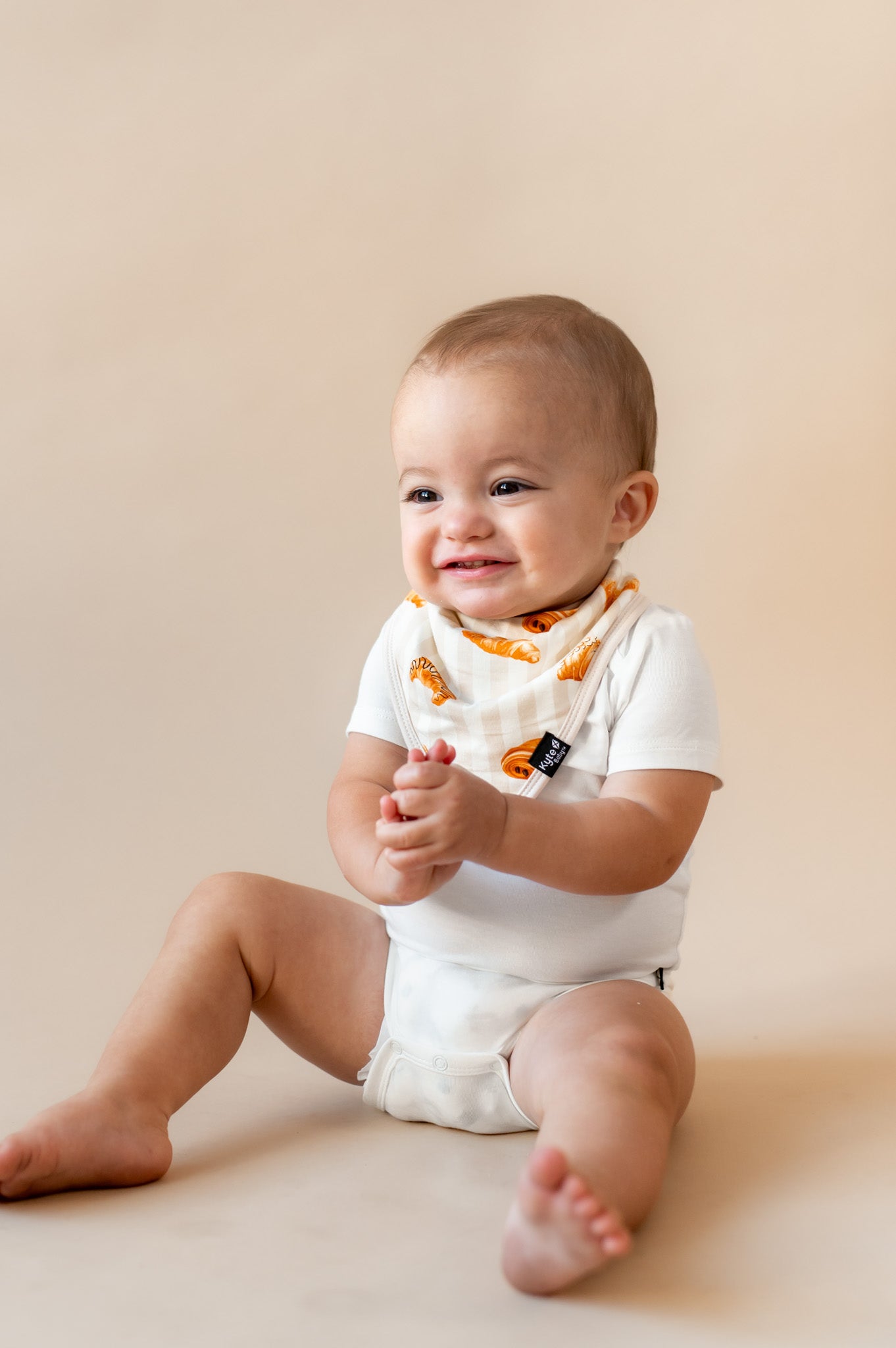 Baby smiling wearing a Bib with Croissant print over white bodysuit on a beige background