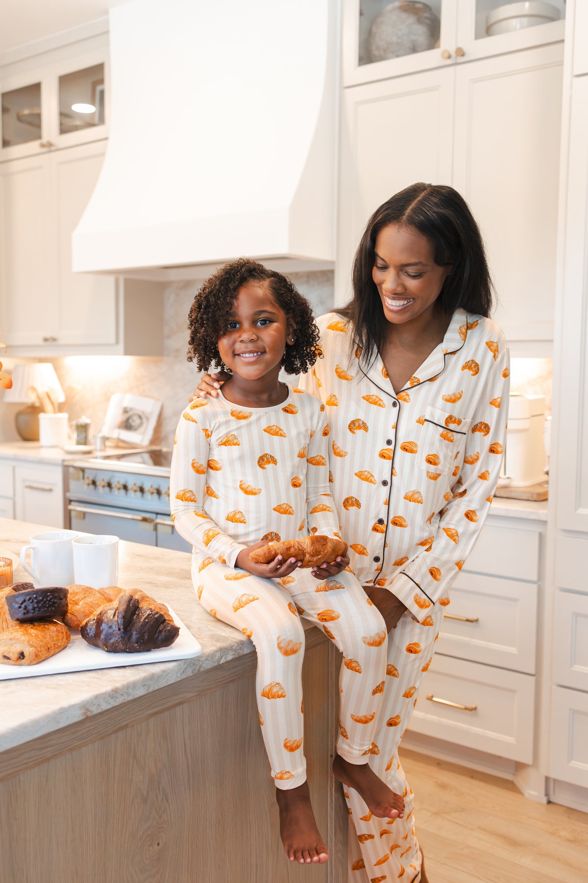 A woman and a young girl wearing matching cream-colored pajamas with a repeating orange and yellow pastry print, sitting together on a kitchen island holding pastries.