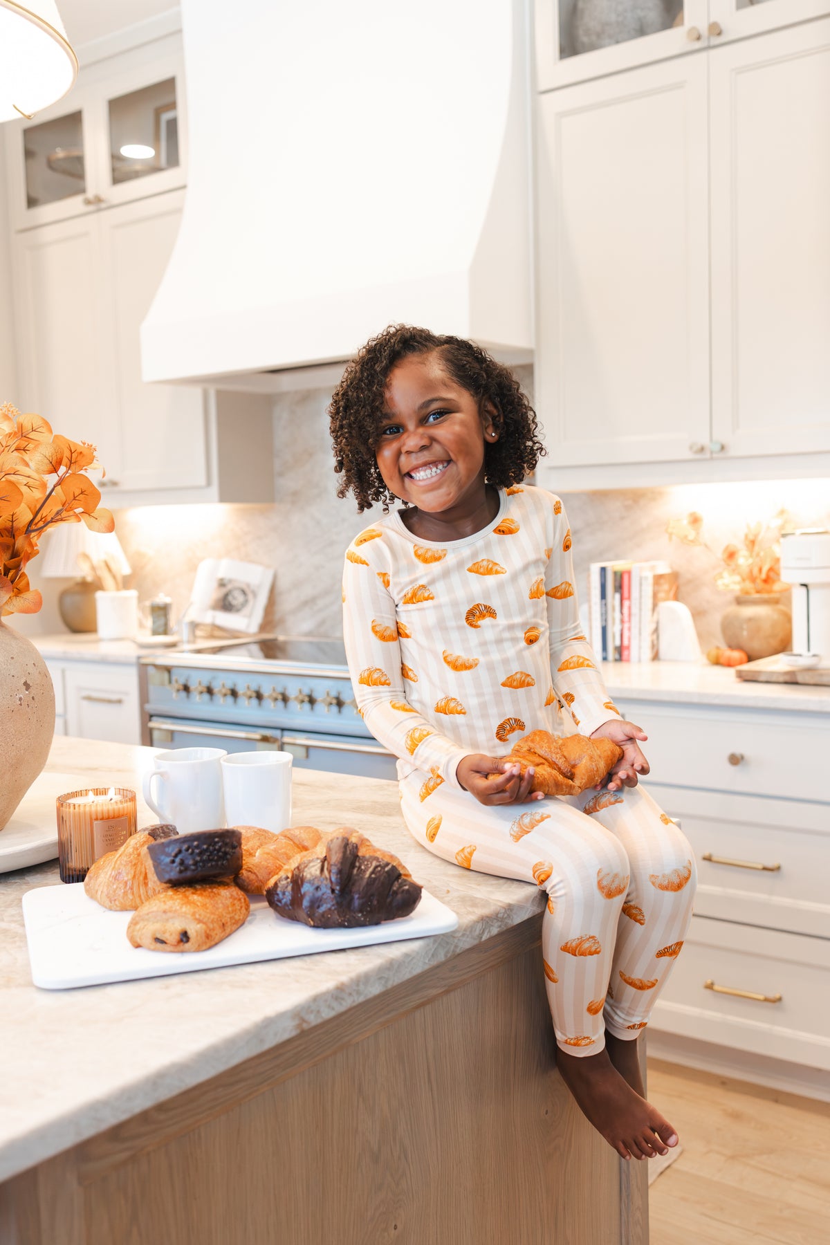 Girl sitting on kitchen counter with croissants wearing Long Sleeve Pajamas with Croissant print holding a croissant