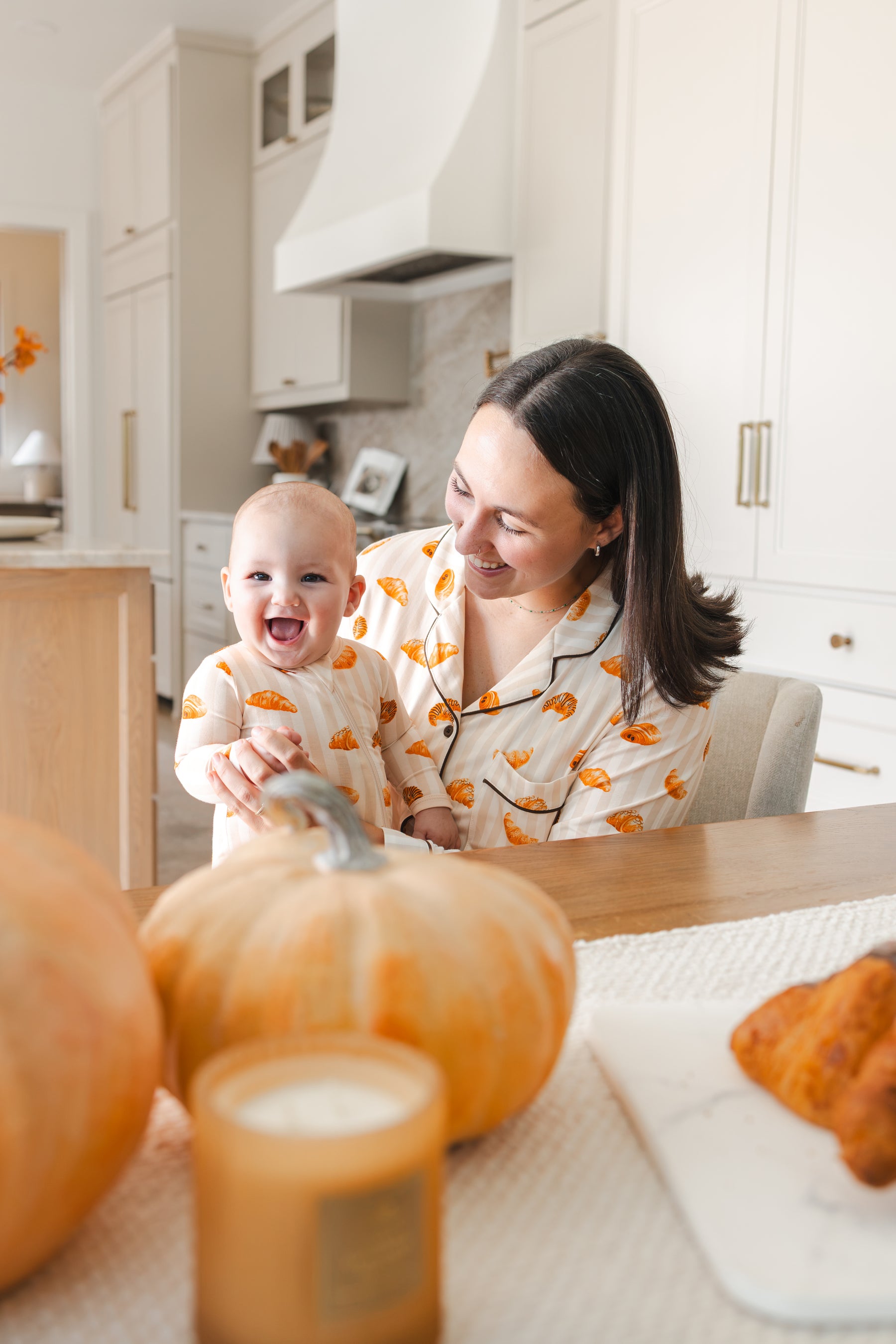 A woman and a baby wearing matching orange and white croissant-patterned pajamas smile and laugh together in a bright, modern kitchen with pumpkins on the table in the foreground.