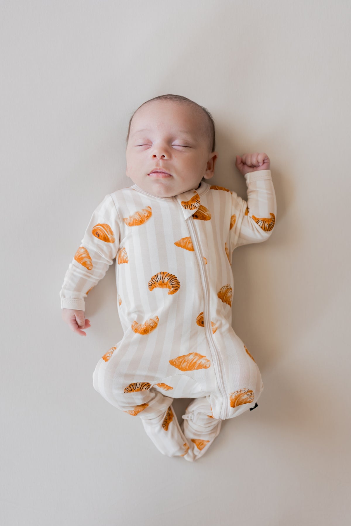 Baby sleeping in Zippered Footie with Croissant print on a beige background