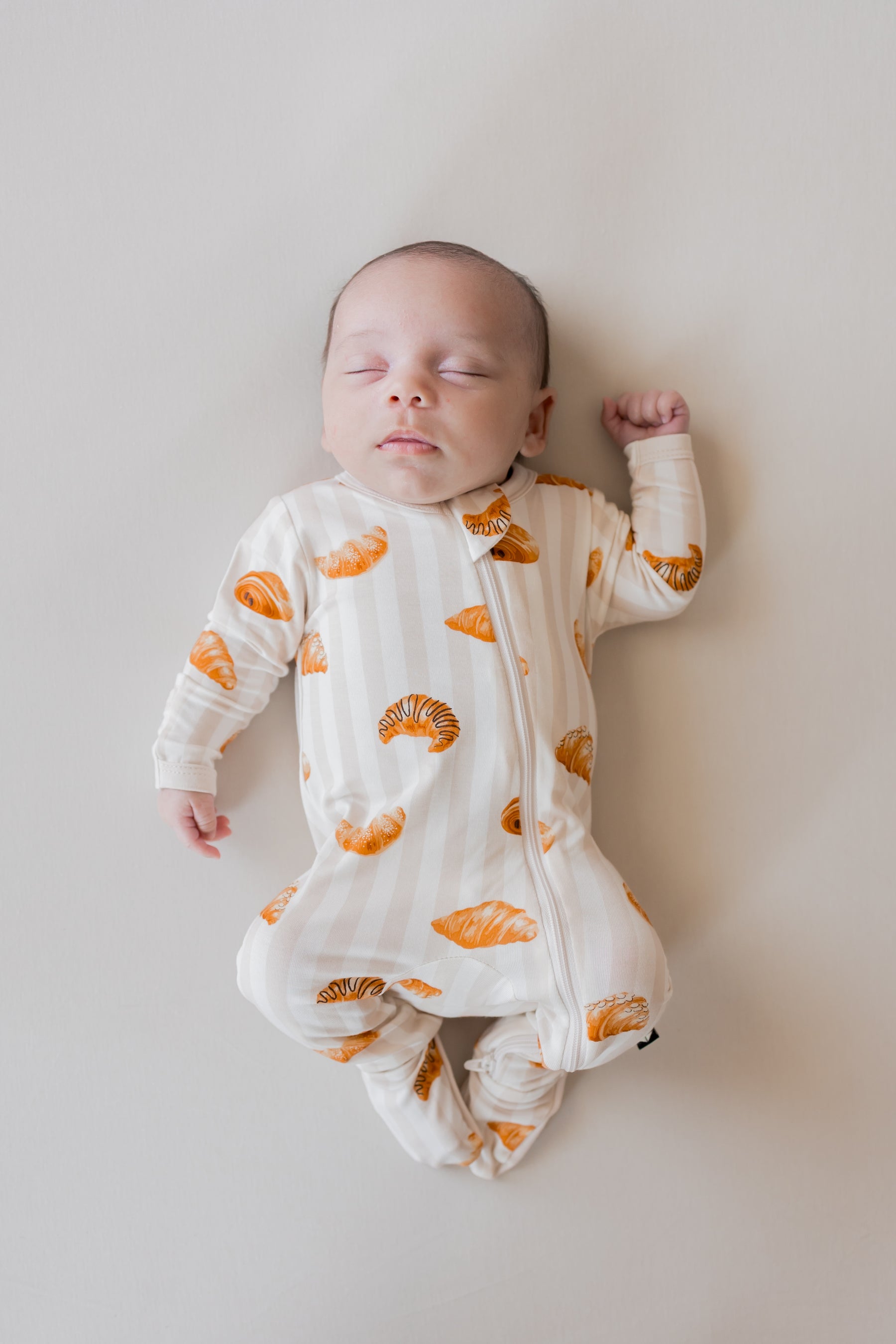 Baby sleeping in Zippered Footie with Croissant print on a beige background