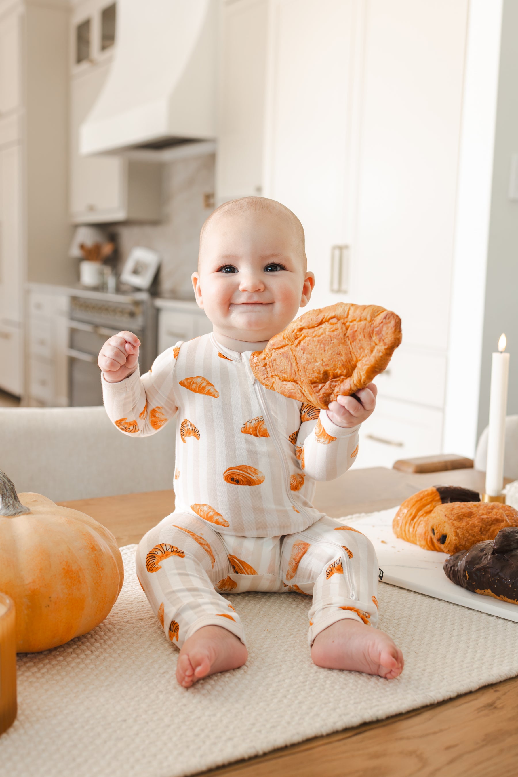 Baby sitting on a table wearing Zippered Romper with Croissant print while holding a croissant