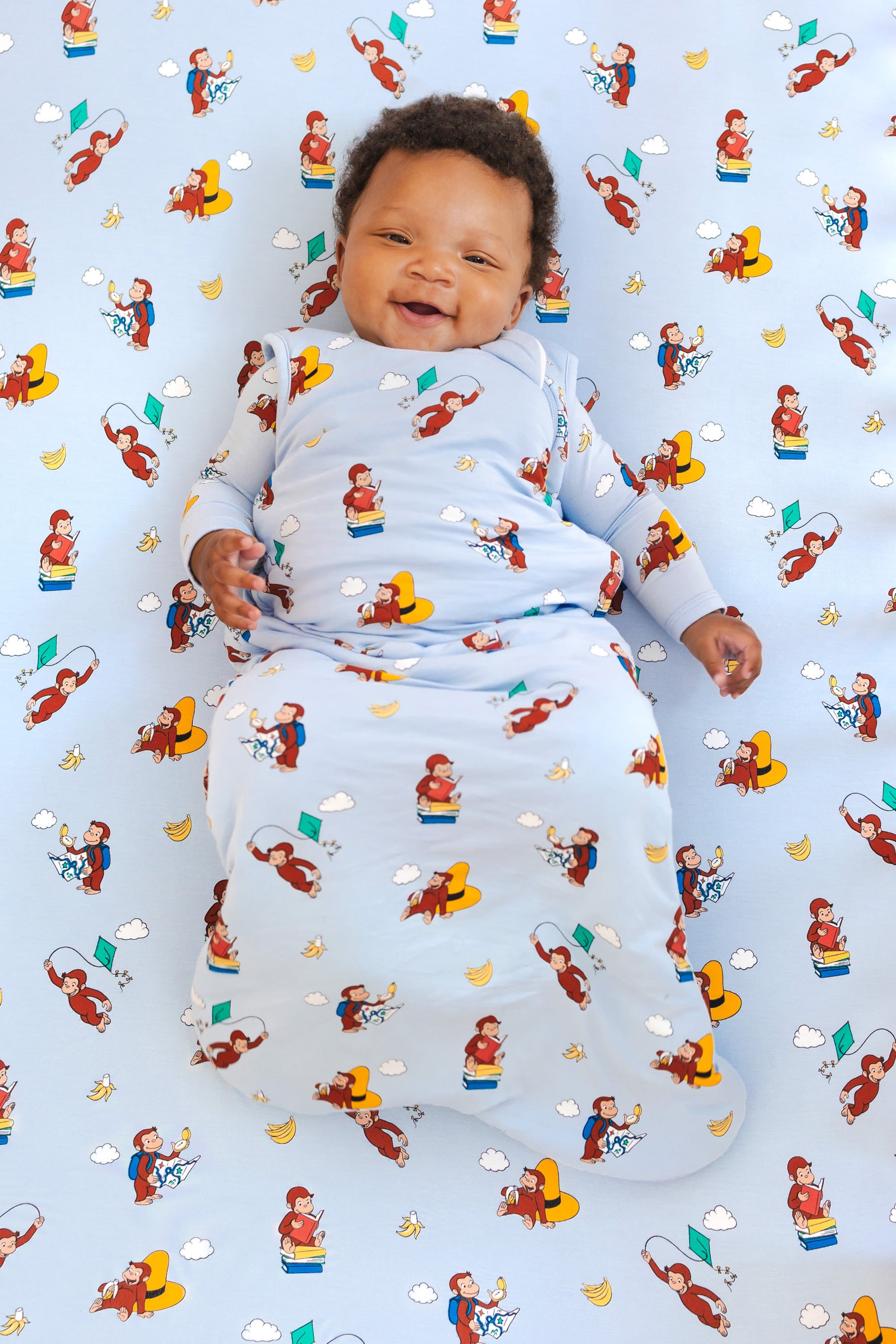 Baby smiling in Sleep Bag in Curious George 1.0 print lying on a similar print crib sheet