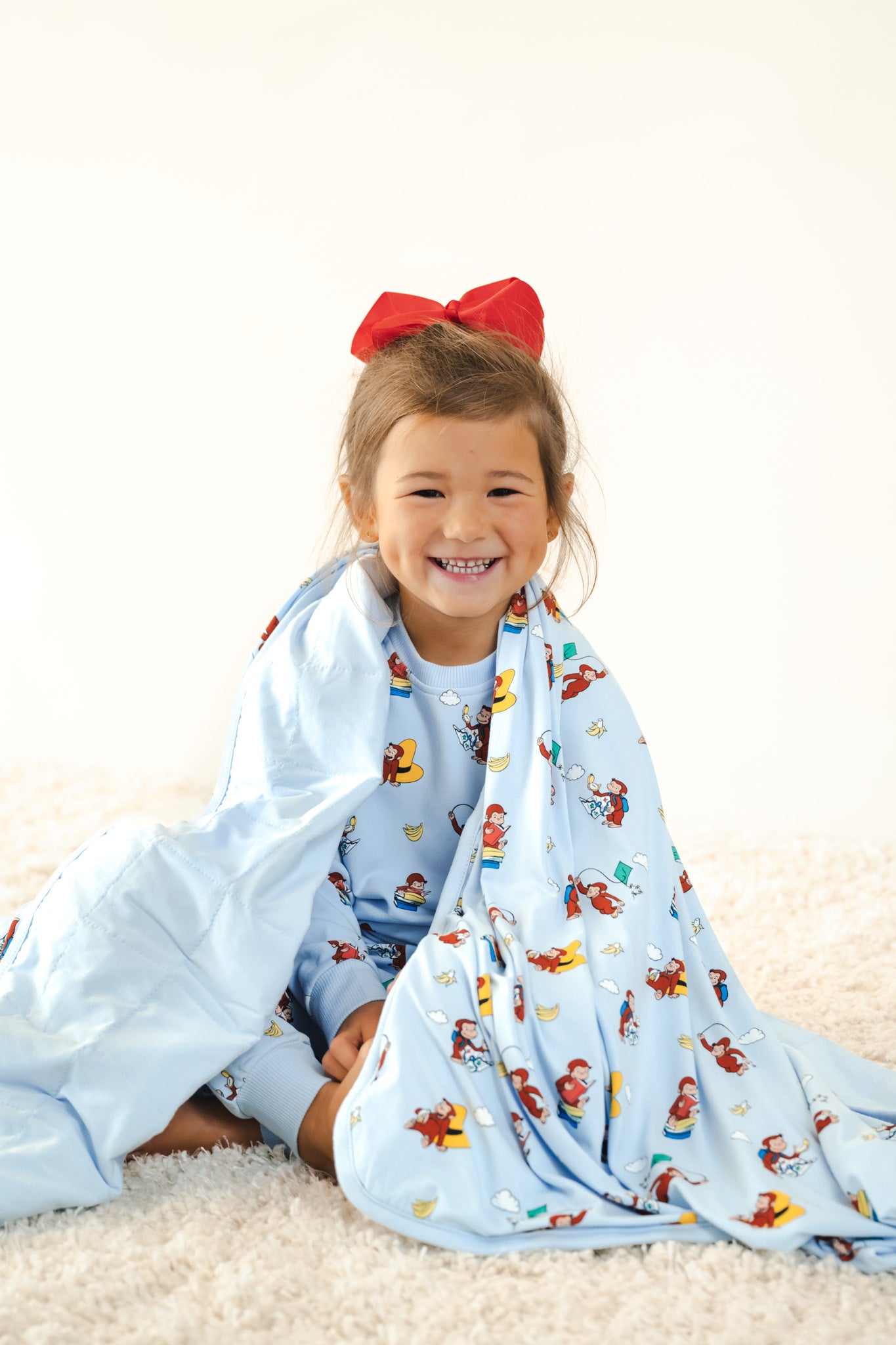 Girl sitting on white carpet wrapped in Toddler Blanket in Curious George 1.0 print wearing similar print long sleeve jogger set and red bow on head