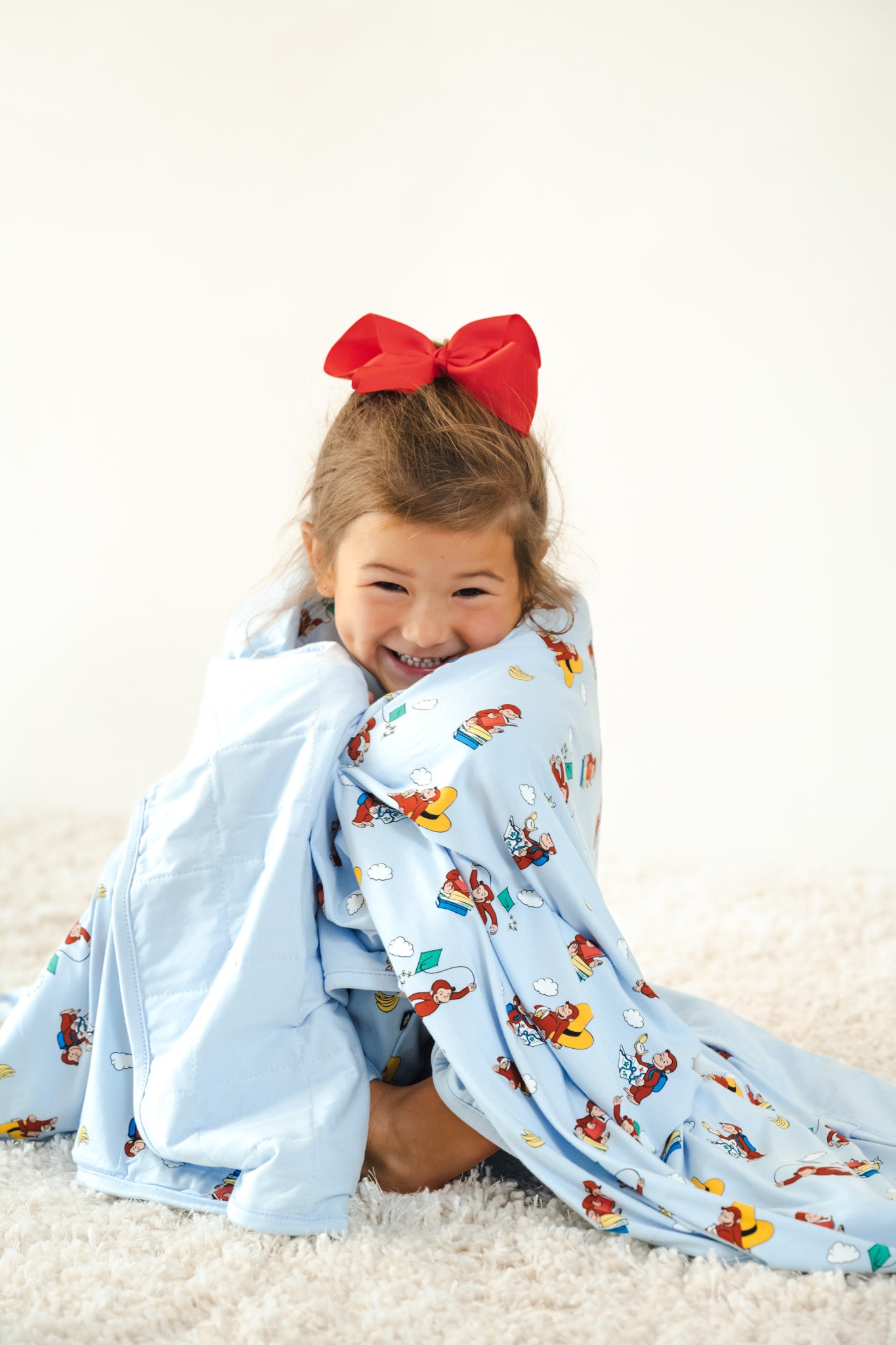 Girl smiling wrapped in Toddler Blanket in Curious George 1.0 print with red bow on head sitting on a white carpet