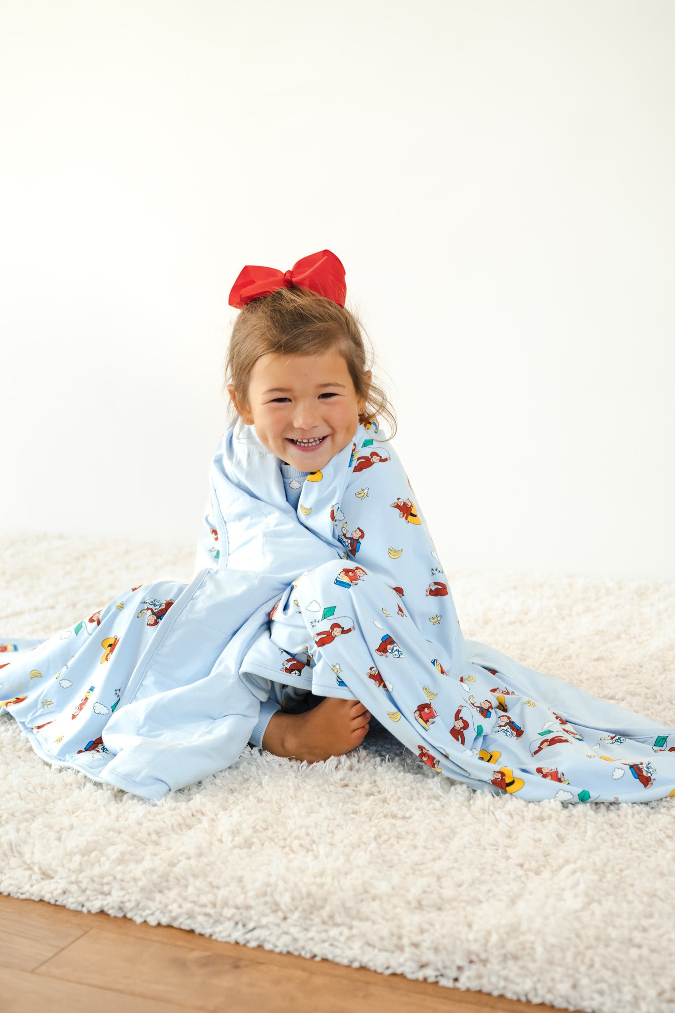 Girl wrapped in Toddler Blanket in Curious George 1.0 print with red bow on head sitting on a white carpet