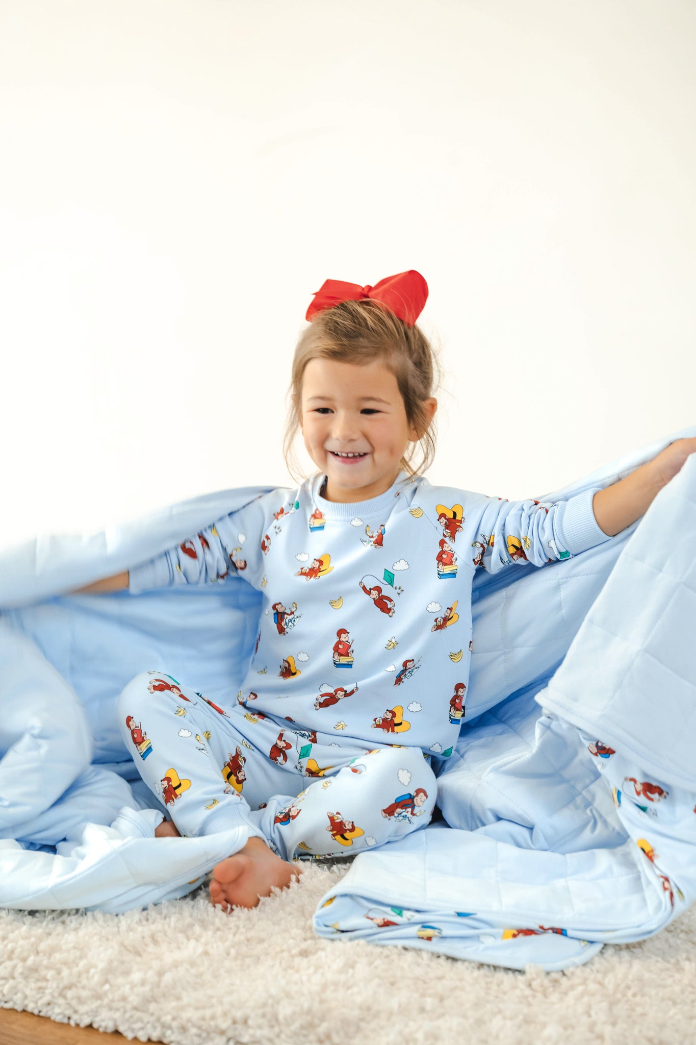 Girl holding Youth Blanket in Curious George 2.5 print wearing similar print long sleeve jogger set and red bow on head sitting on a white carpet