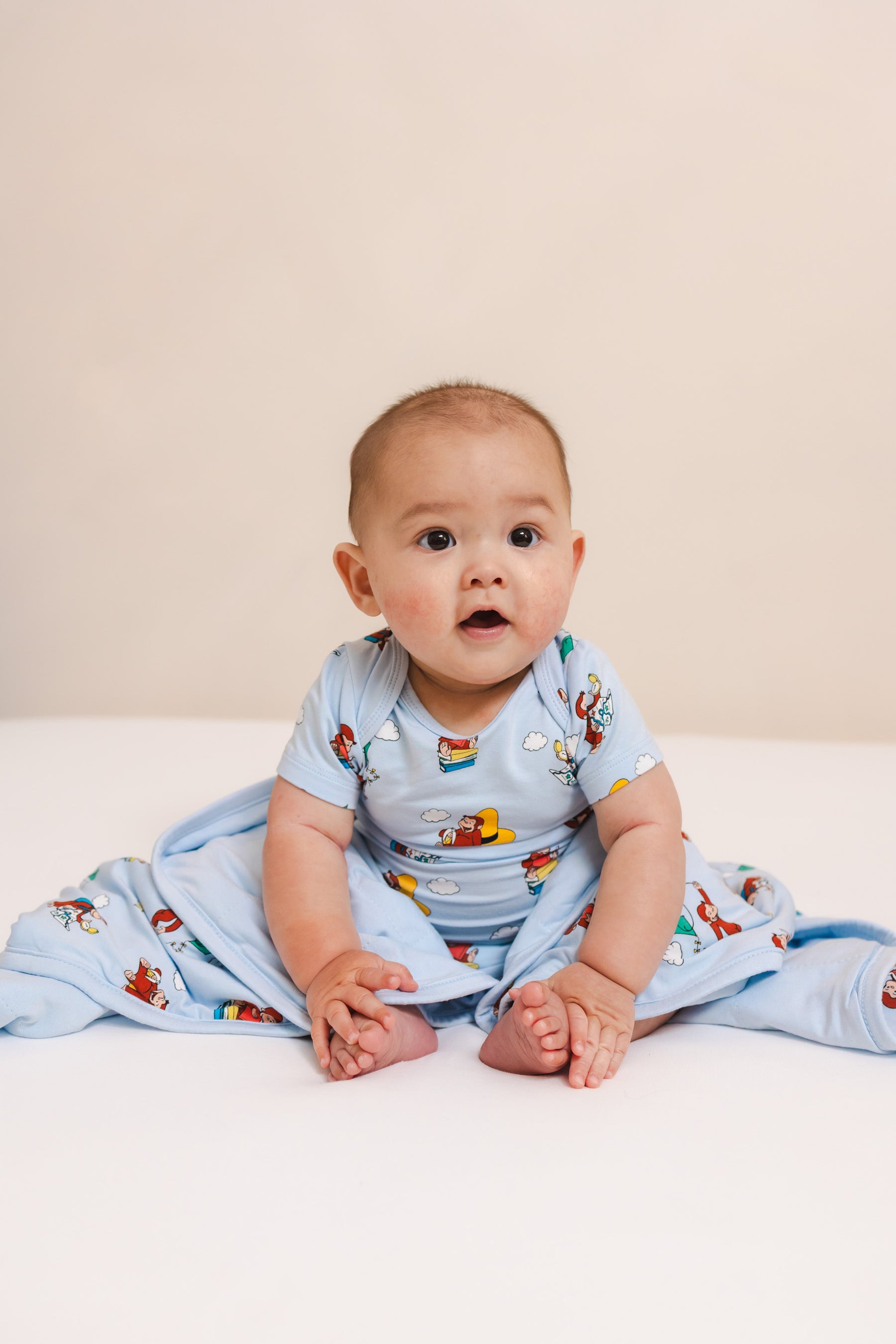 Baby wrapped in Curious George 1.0 print Baby Blanket wearing similar print bodysuit on a beige background