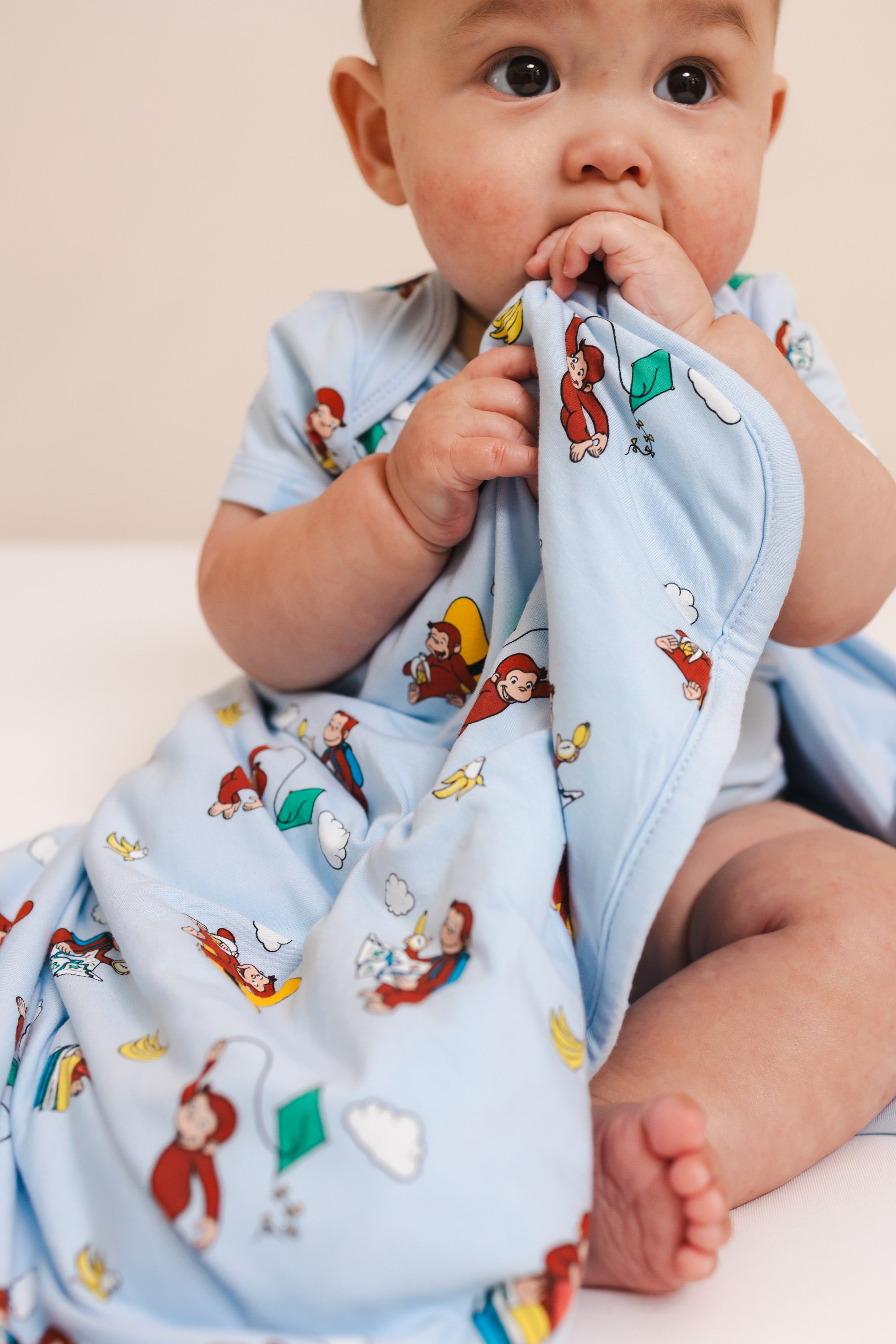 Baby sitting with Baby Blanket in Curious George 1.0 print wearing similar print bodysuit on a beige background