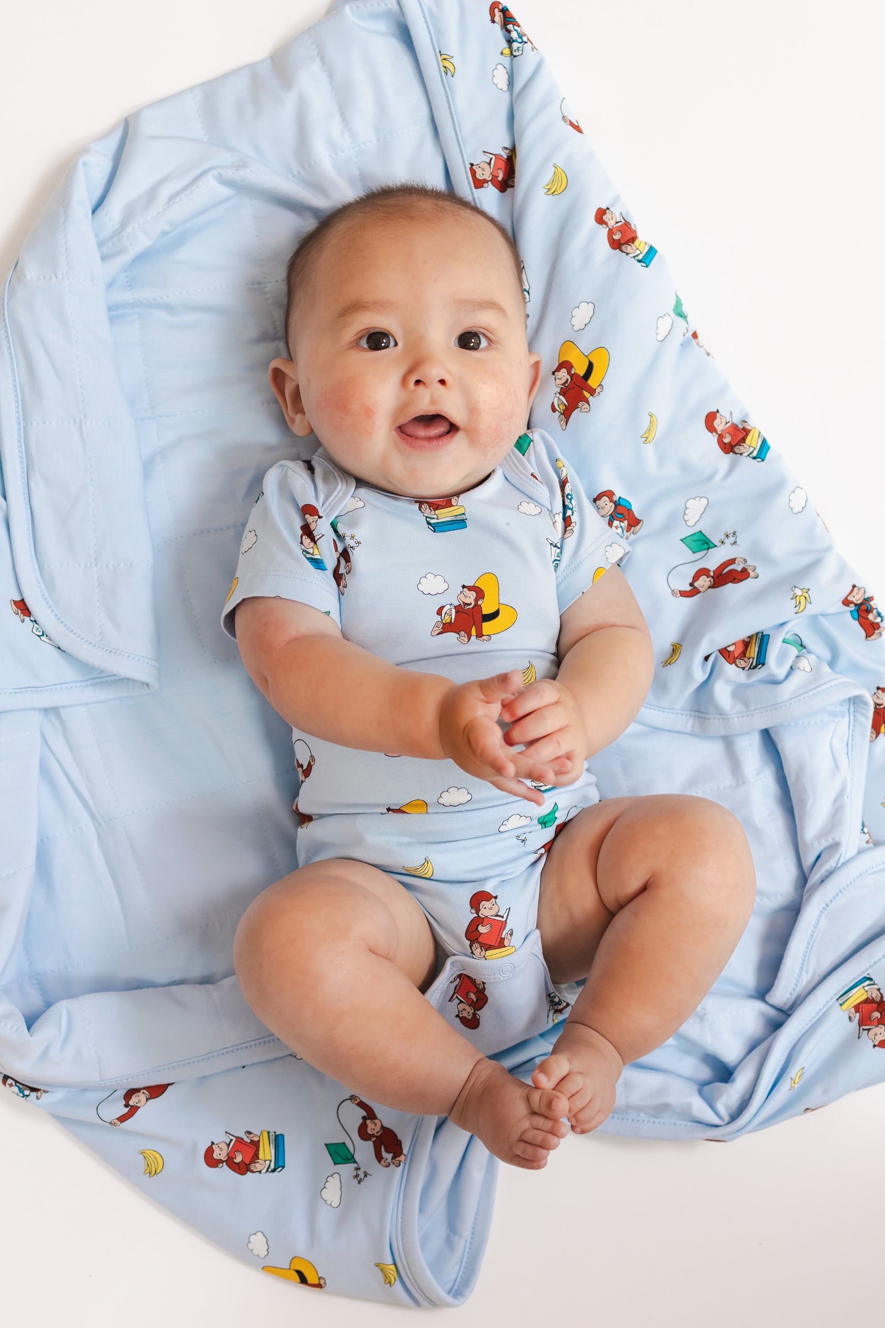 Baby lying on Baby Blanket in Curious George 1.0 print wearing similar print bodysuit on a beige background