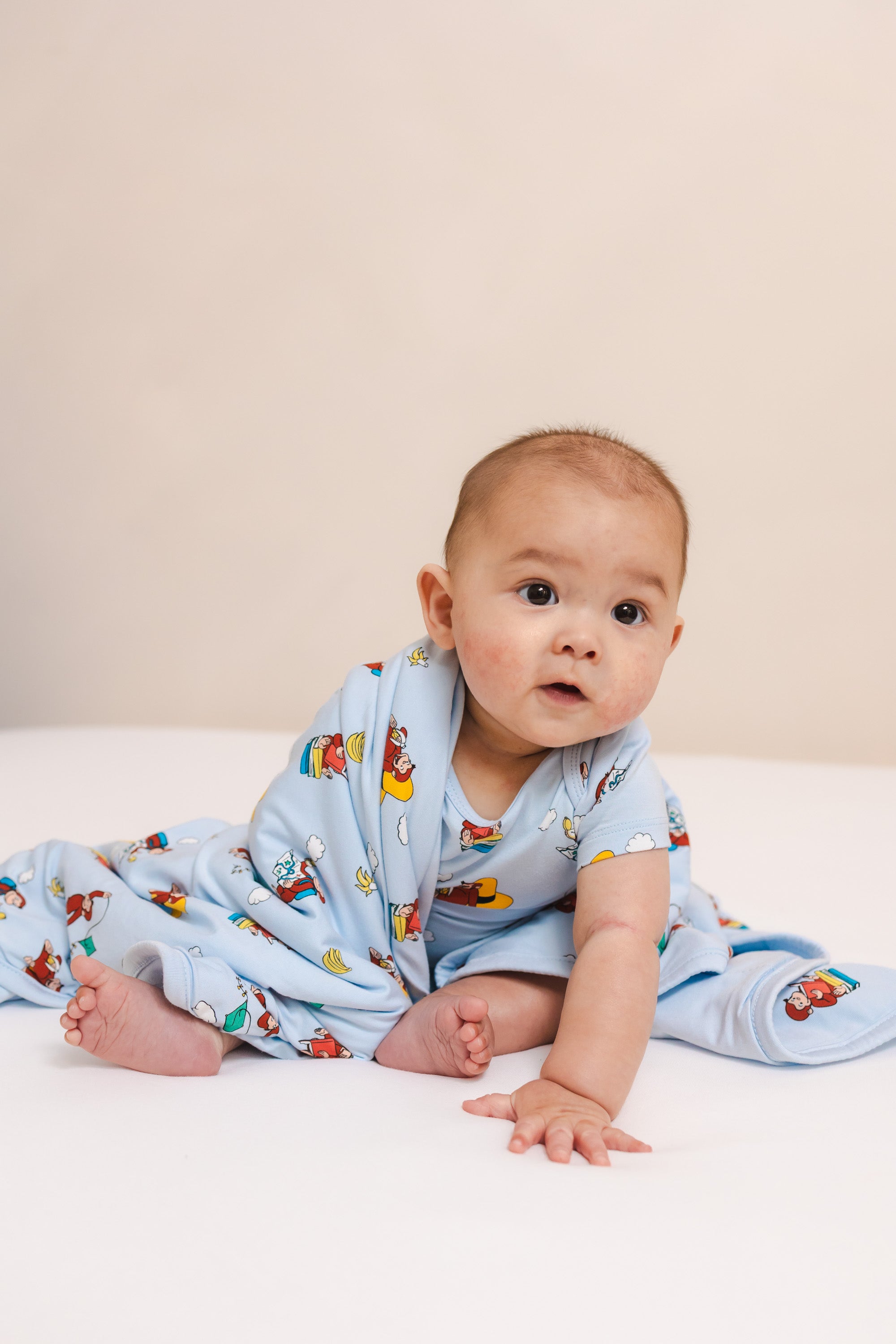 Baby sitting wrapped in Baby Blanket in Curious George 1.0 print wearing similar print bodysuit on a beige background