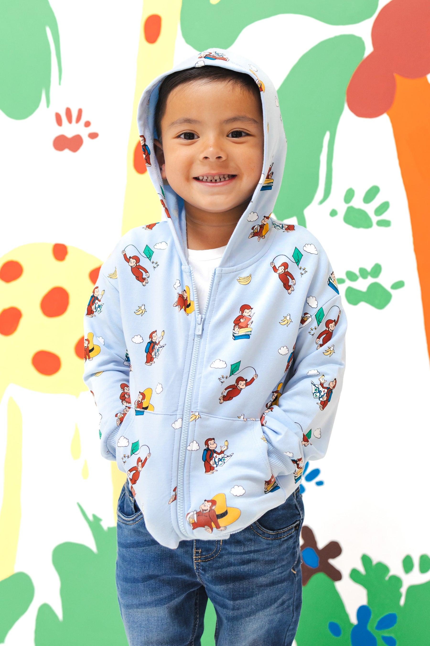 Boy wearing Zip-Up Hoodie in Curious George print over a blue jeans standing in front of a colorful background