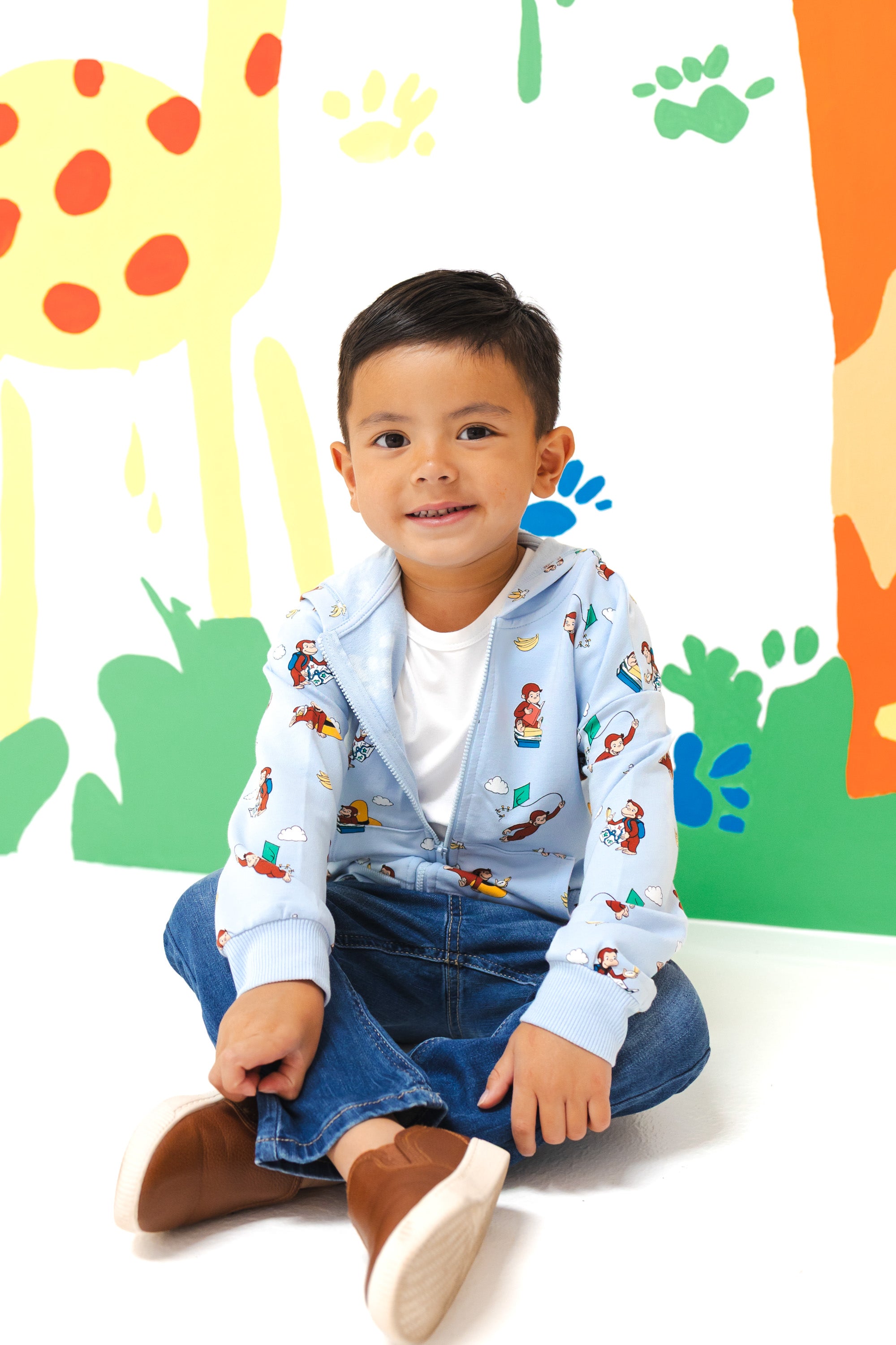 Boy sitting wearing Zip-Up Hoodie in Curious George print over a blue jeans in front of a colorful background