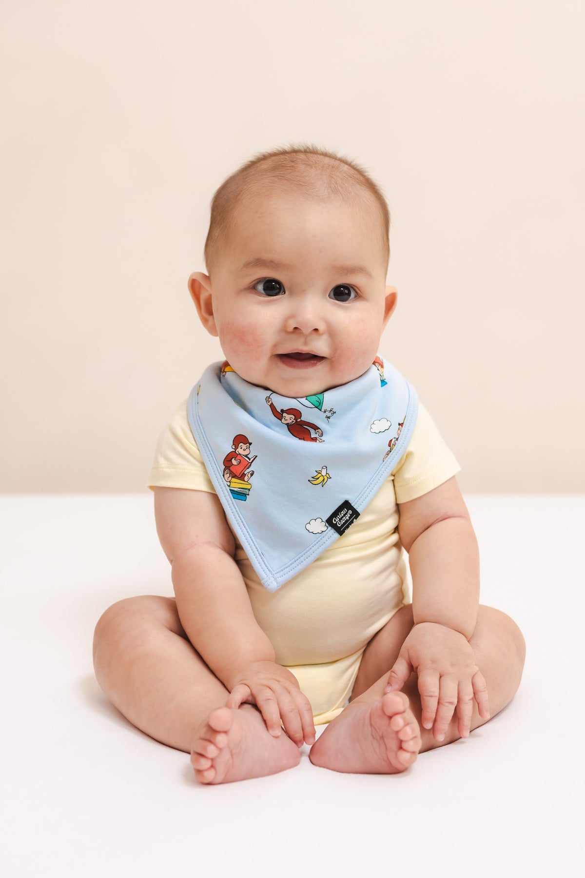 Baby wearing a Bib in Curious George print over a yellow bodysuit sitting on a beige background