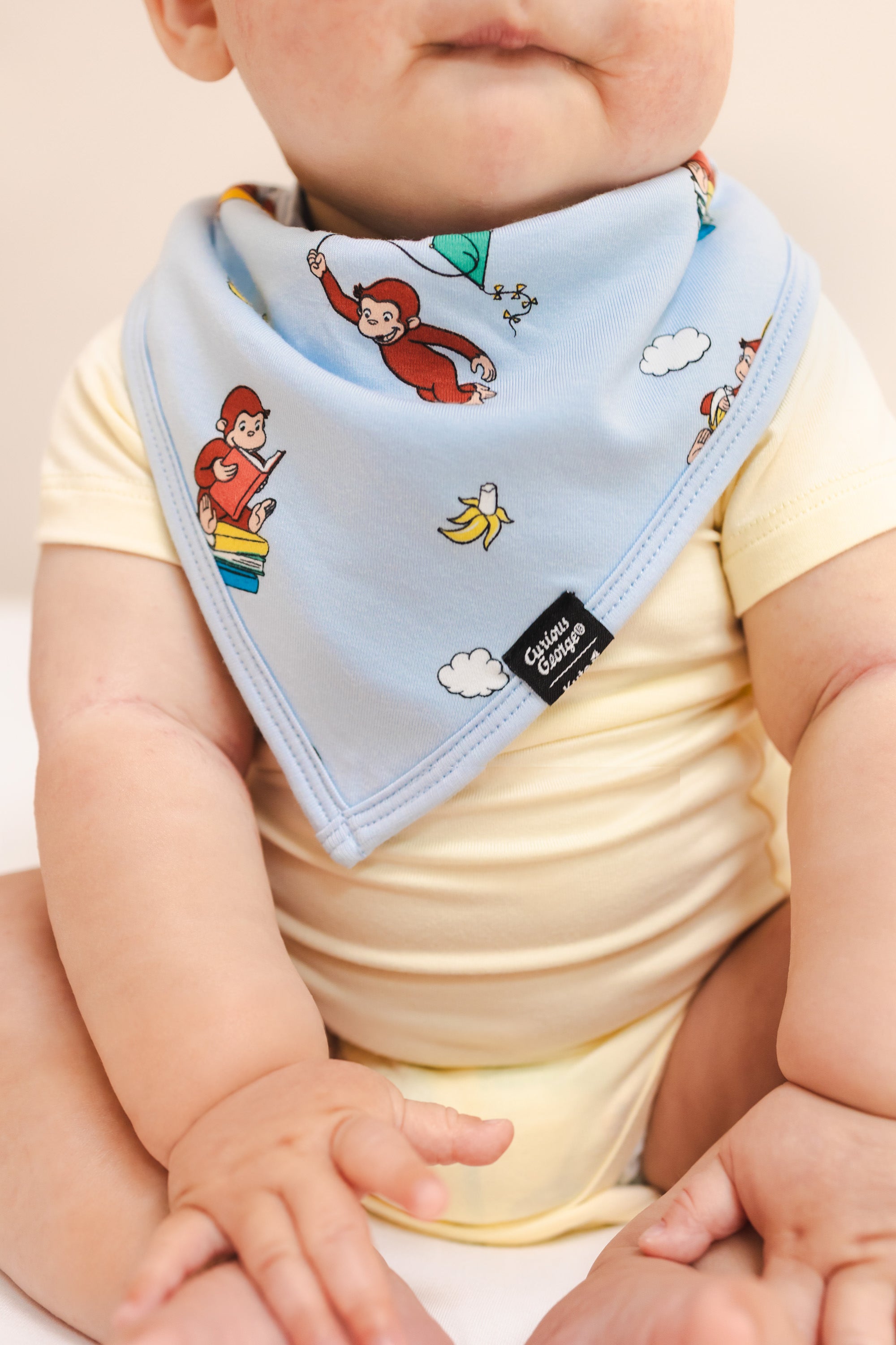 Close up of Baby wearing a Bib in Curious George print over a yellow bodysuit sitting on a beige backgrounds George