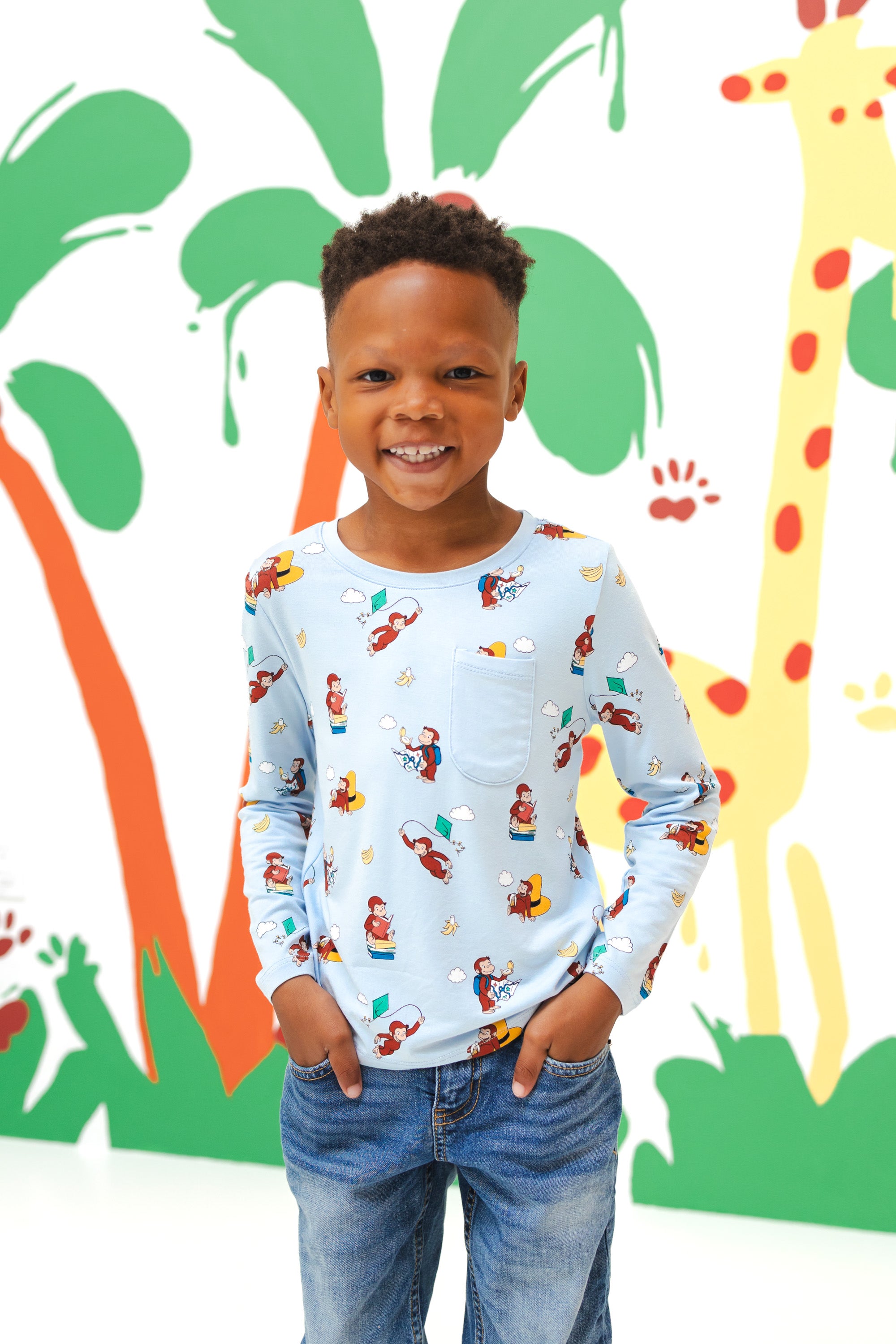 Boy standing in front of a colorful background wearing Long Sleeve Toddler Crew Neck Tee in Curious George print over a blue jeans