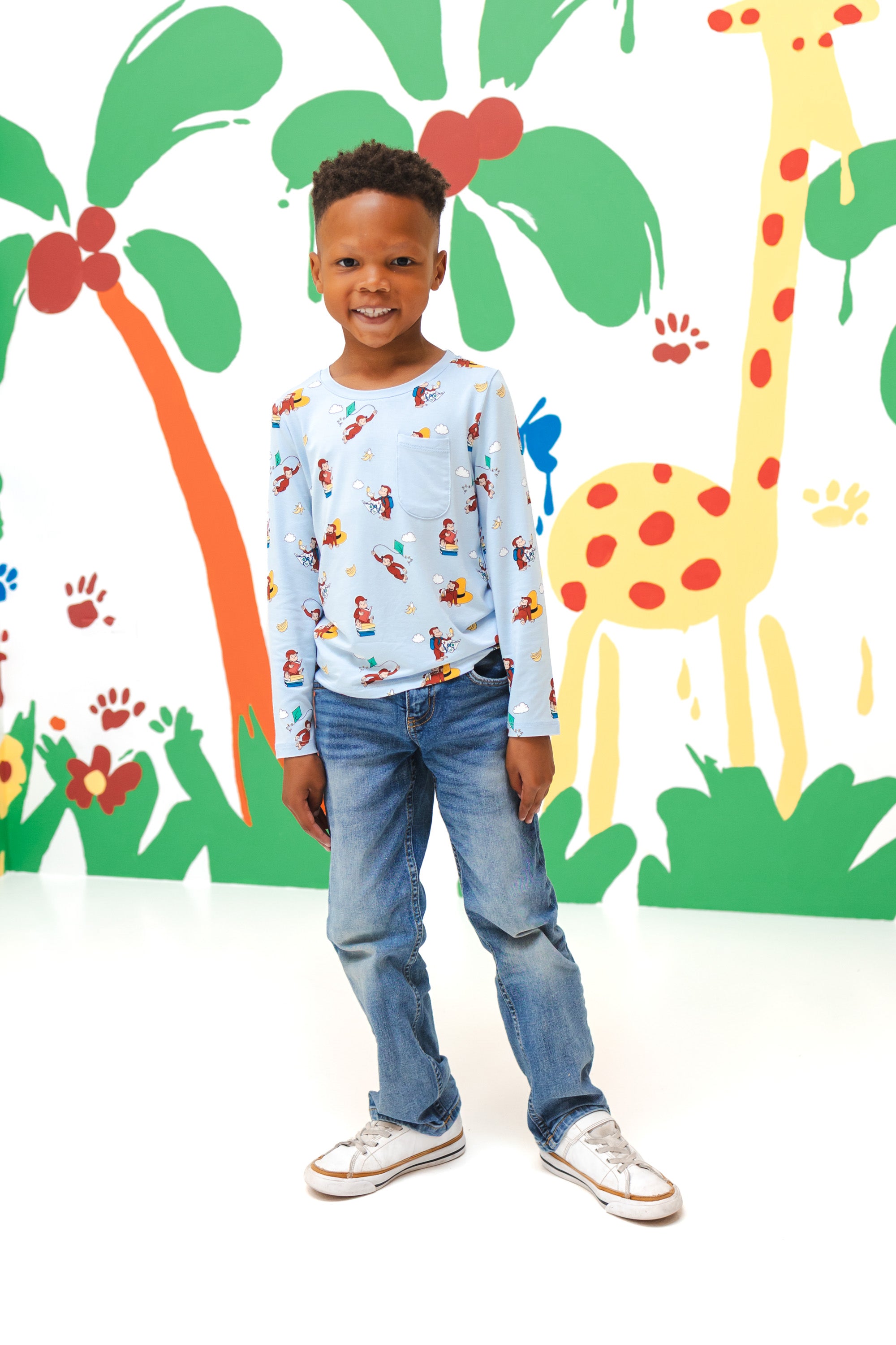 Boy posing in front of a colorful background wearing Long Sleeve Toddler Crew Neck Tee in Curious George print over a blue jeans