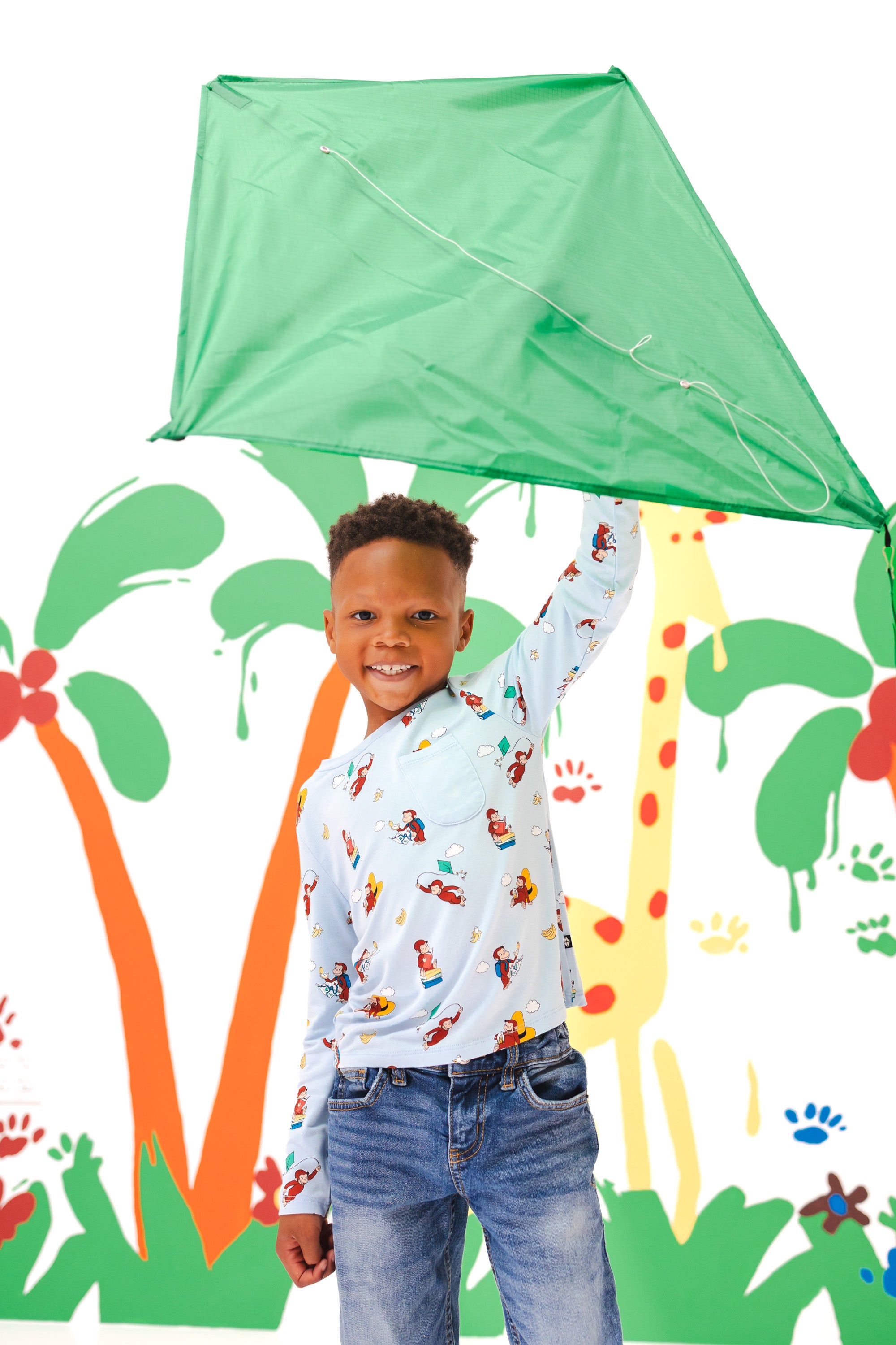 Boy holding a kite standing in front of a colorful background wearing Long Sleeve Toddler Crew Neck Tee in Curious George print over a blue jeans