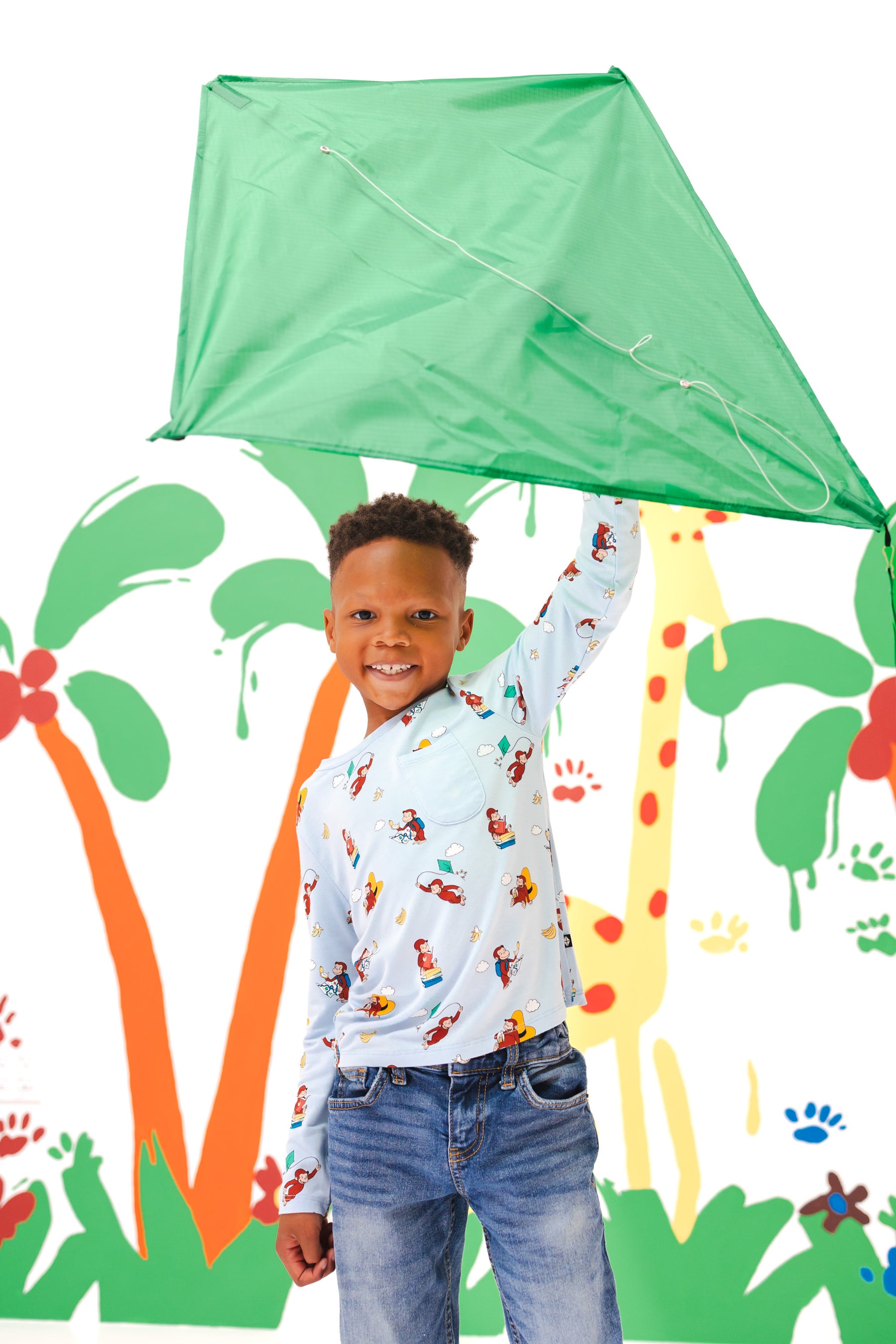 Boy holding a kite standing in front of a colorful background wearing Long Sleeve Toddler Crew Neck Tee in Curious George print over a blue jeans