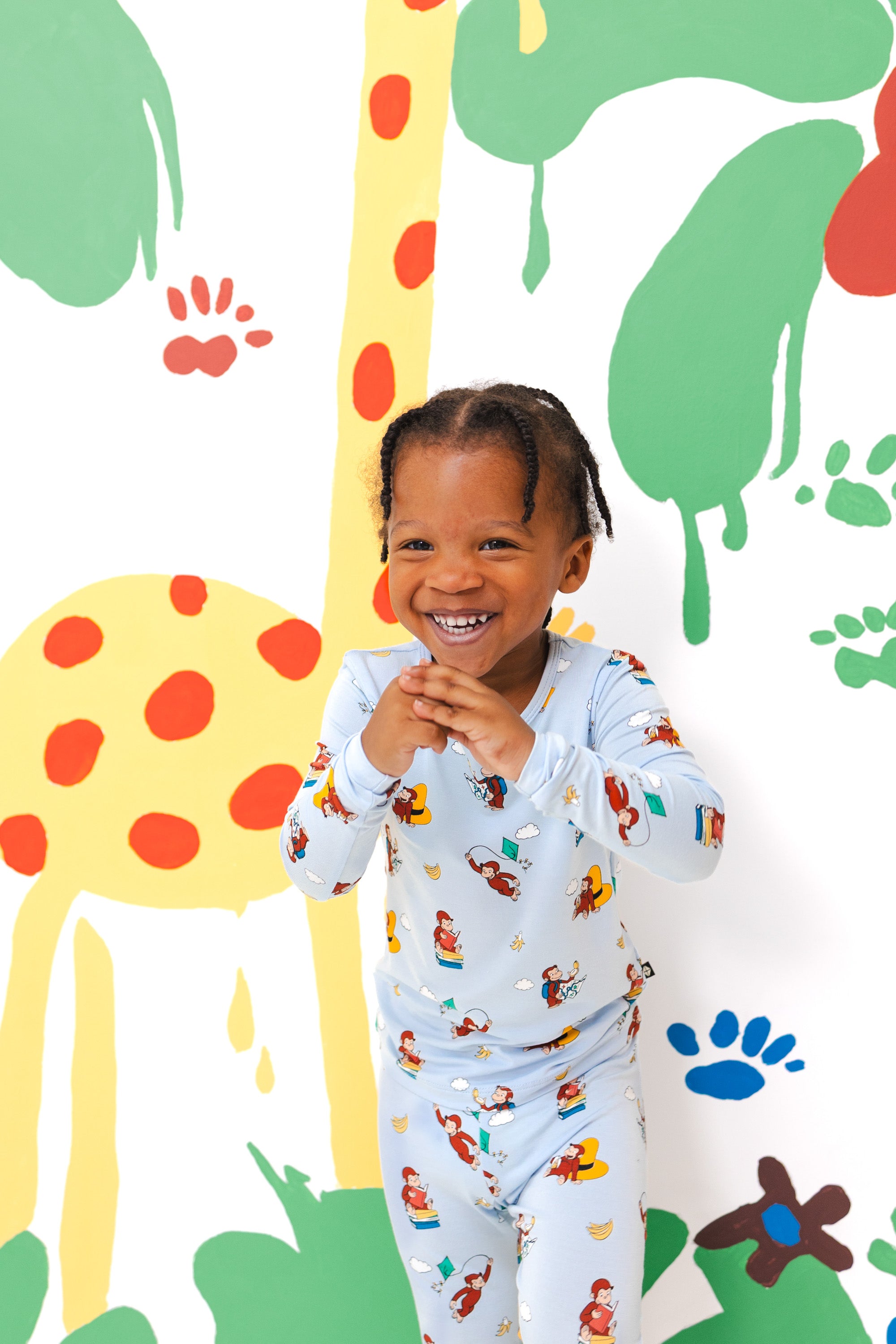 Girl smiling in Long Sleeve Pajamas in Curious George print standing in front of a colorful background