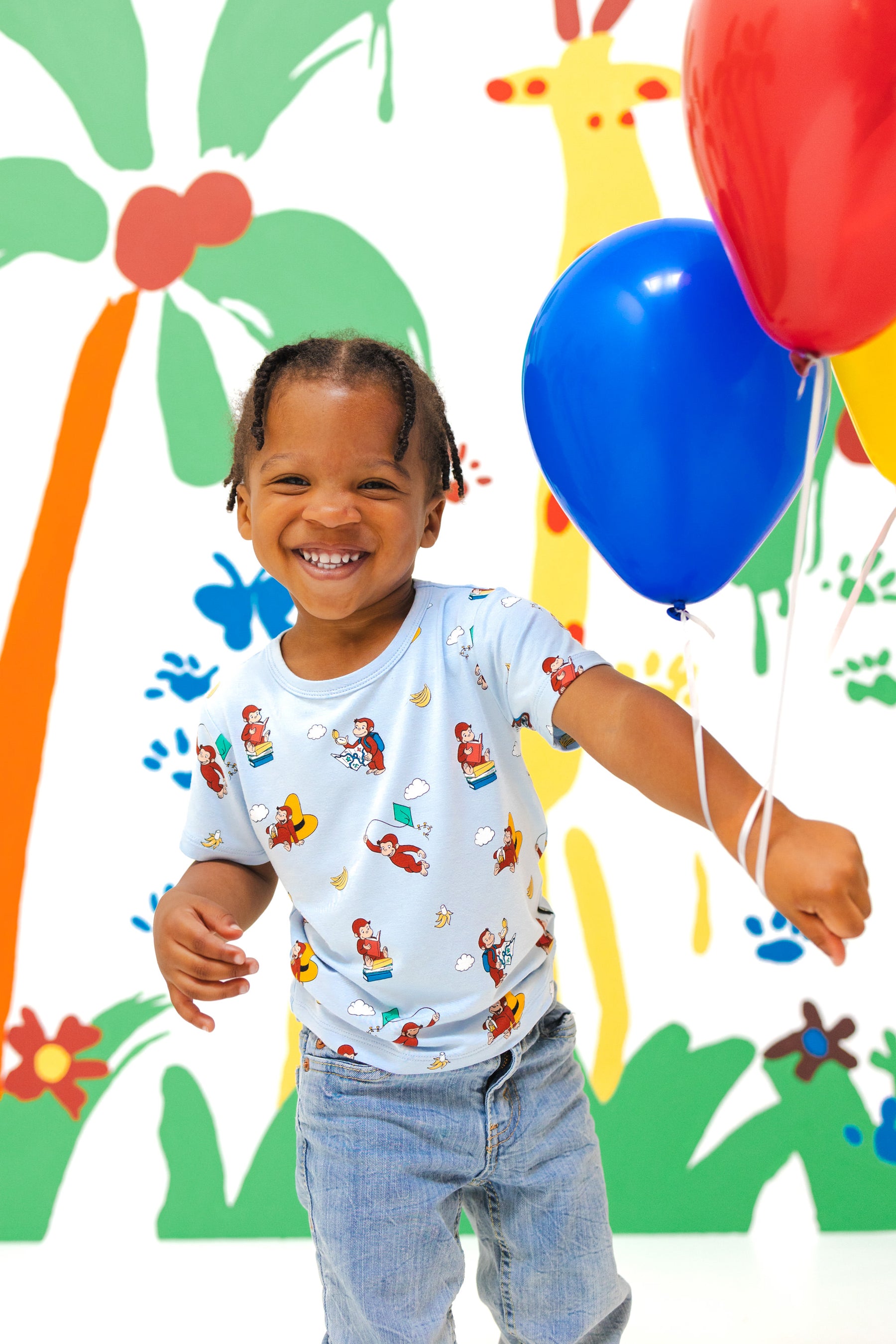 Girl wearing Toddler Basic Tee in Curious George print over a blue jeans holding red blue balloons in front of a colorful background