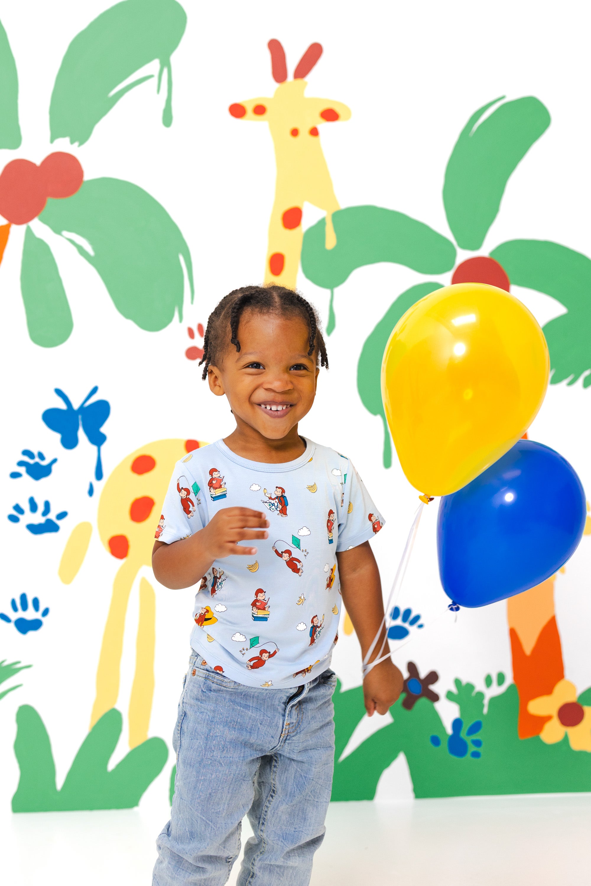 Girl holding red blue balloons wearing Toddler Basic Tee in Curious George print over a blue jeans smiling in front of a colorful background