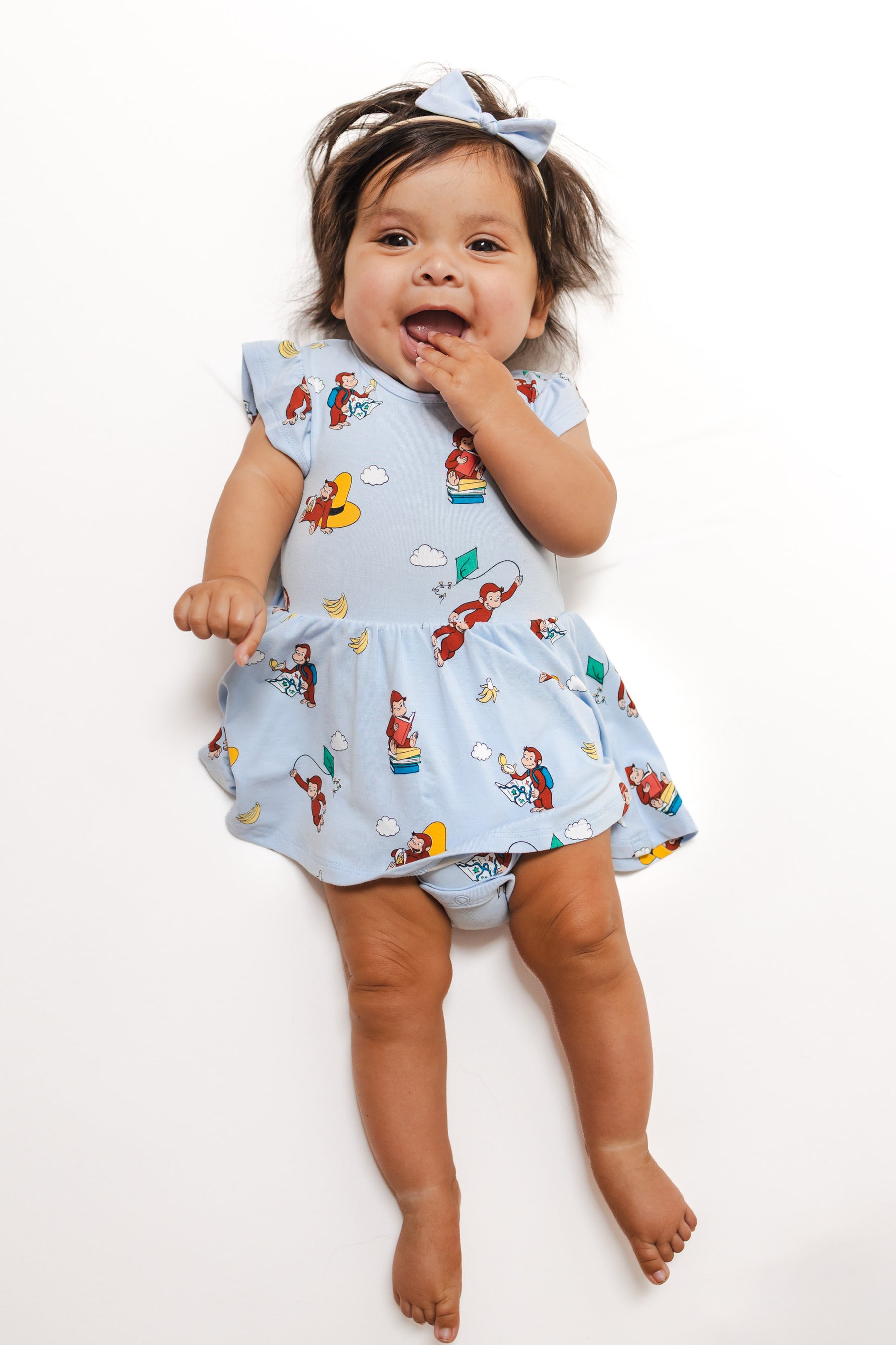 Baby wearing Twirl Bodysuit Dress in Curious George print on a white background with a bow tie on head