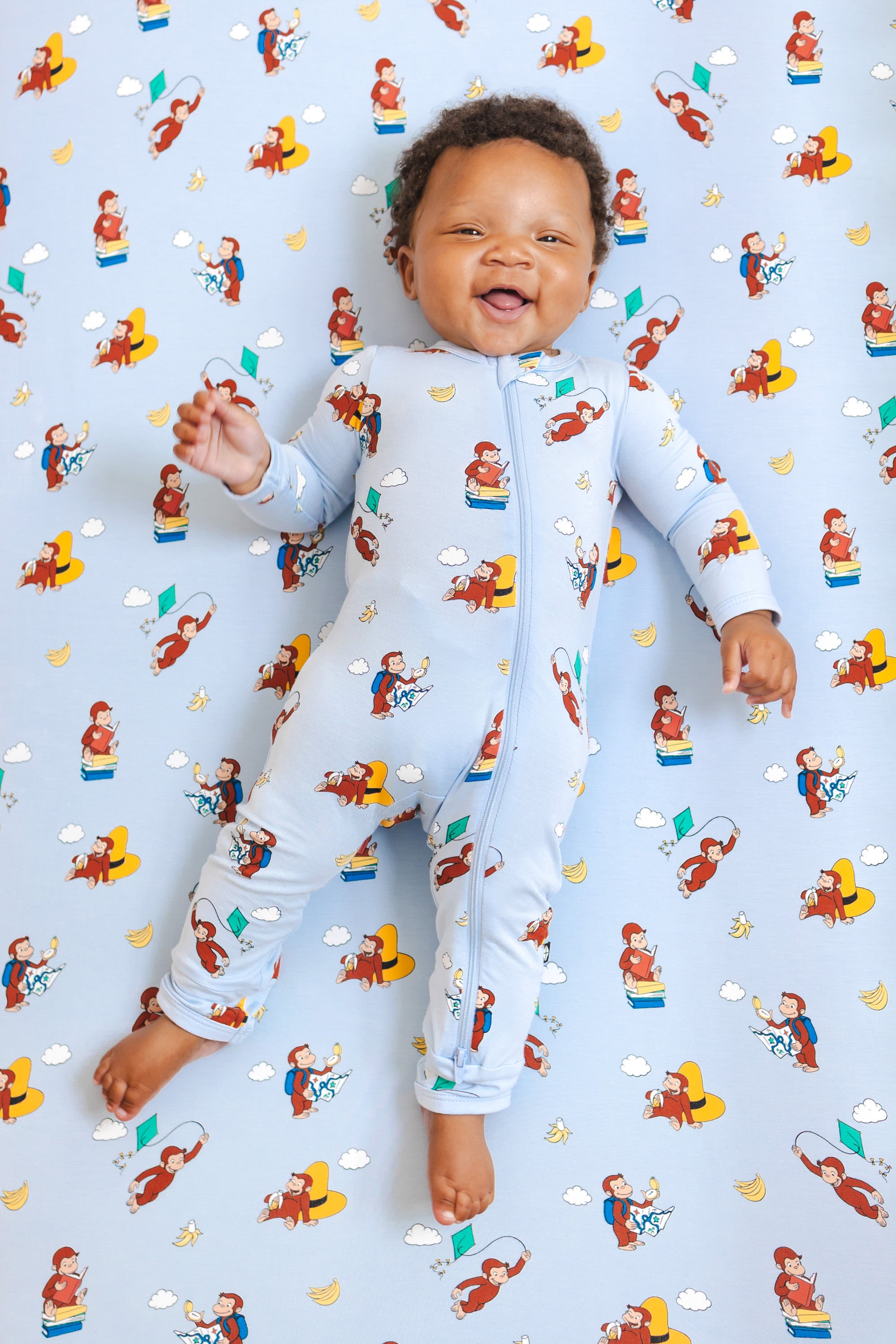 Baby smiling in Zippered Romper in Curious George print lying over a similar print crib sheet