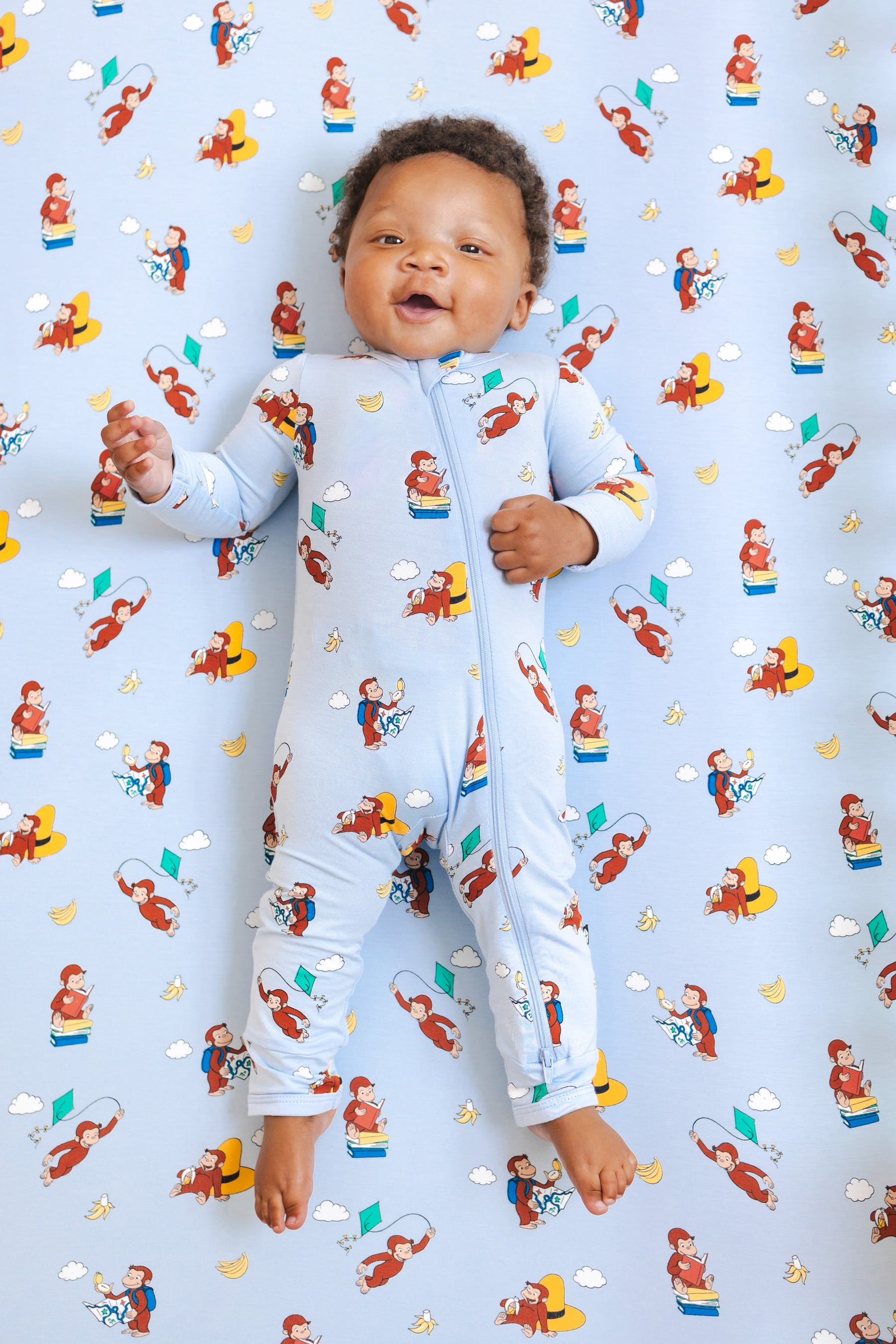 Baby wearing Zippered Romper in Curious George print lying over a similar print crib sheet